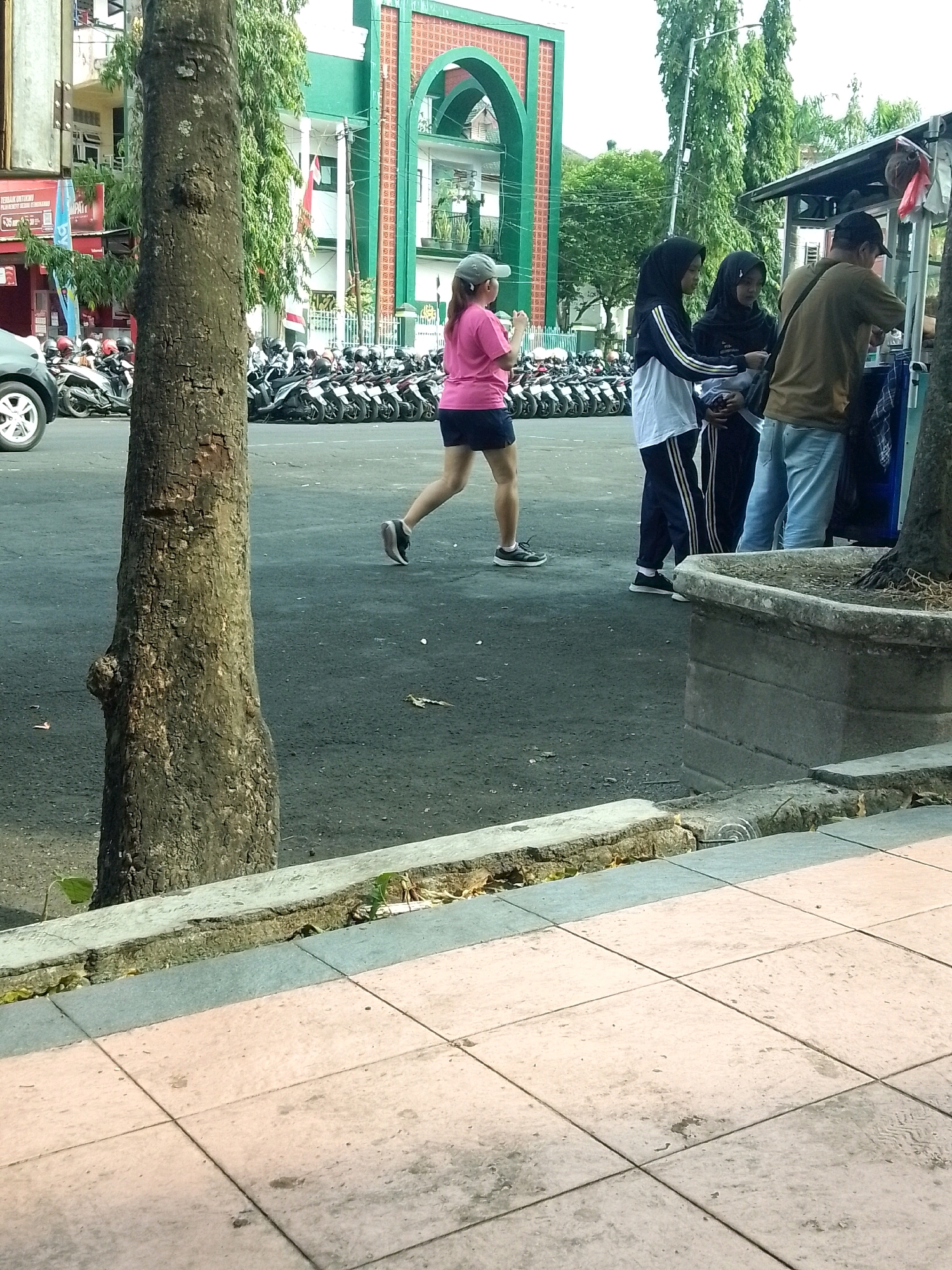 In Jember, Indonesia, a street scene unfolds with a paved road and sidewalk. A large tree trunk stands prominently in the foreground on the left. On the right, a concrete planter box is visible. In the mid-ground, a woman in a pink shirt and dark shorts is walking, wearing a cap. She is facing left, away from the camera. Behind her, a group of three people stand near a food cart. Two individuals, possibly female, are wearing hijabs. The person on the far right is wearing a brown shirt and light blue jeans. Behind this group, a row of parked motorcycles stretches across the scene. In the background, a building with green arched entrances and white architectural details is visible. Trees with green foliage are also present in the background. The overall lighting suggests it is daytime.