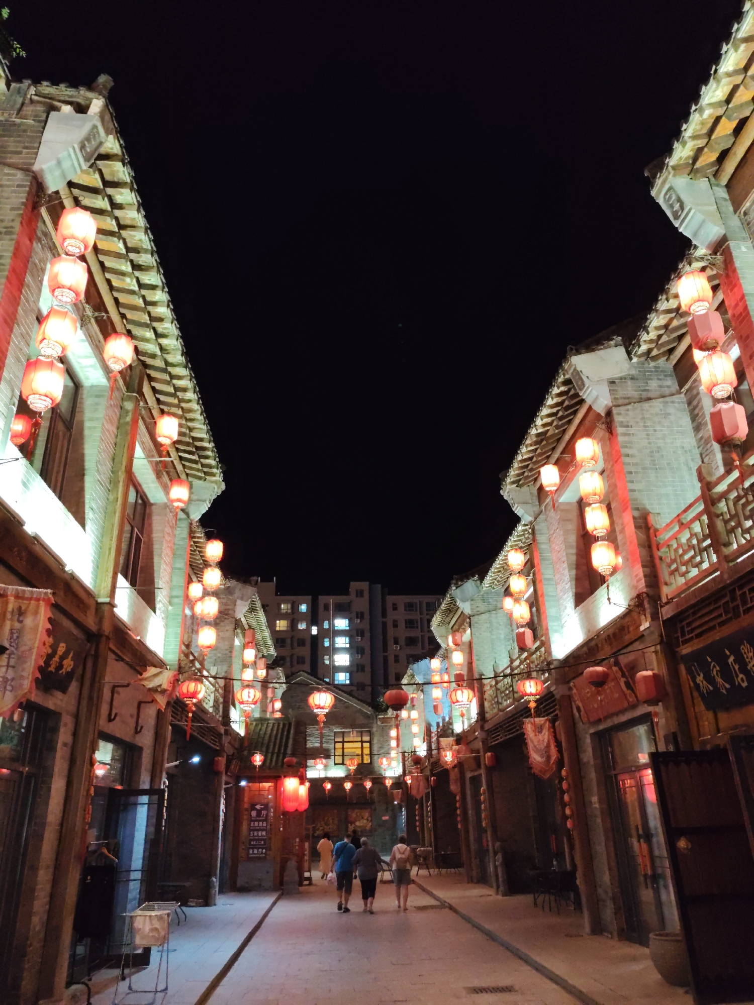 This is a nighttime photograph of a street in Lindian, China, decorated with numerous red lanterns that illuminate the traditional architecture. The street is paved and lined with buildings that feature tiered, tiled roofs and brick facades, some with carved wooden elements. The lanterns, predominantly round and cylindrical, hang from wires strung across the street and along the building fronts, casting a warm, ambient glow. In the background, a modern, multi-story building with illuminated windows is visible against the dark night sky, contrasting with the older structures. Several people are walking down the street, appearing to be tourists or residents enjoying the evening. The scene conveys a festive and historic atmosphere.
