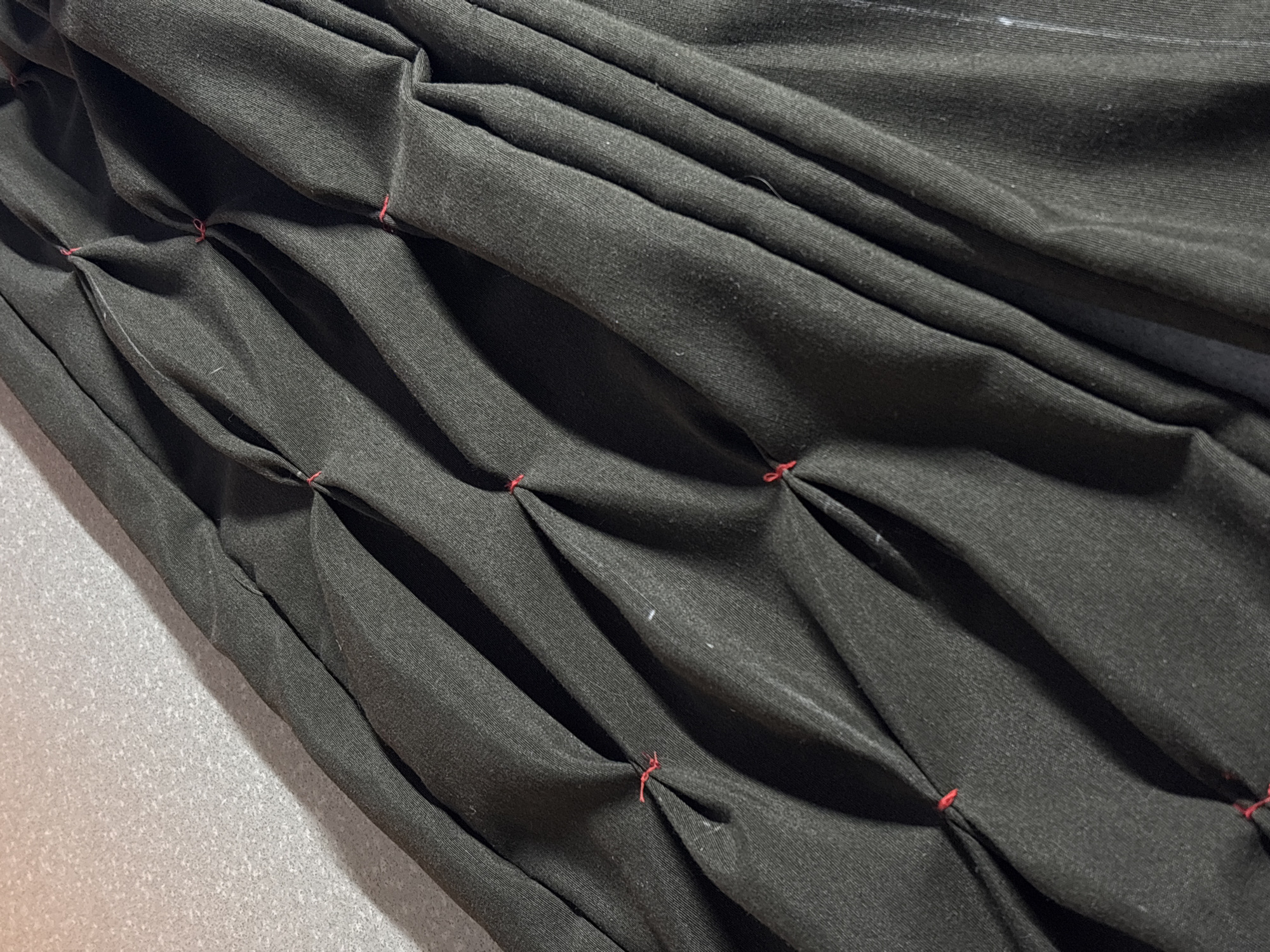The image shows a section of black fabric with a series of pintucks running diagonally across the frame. The pintucks are secured at regular intervals with red thread. The fabric appears to be lying on a gray surface. The scene is likely indoors, but the location is not possible to determine from the given information. No people, objects other than fabric and thread, actions, or interactions are visible.