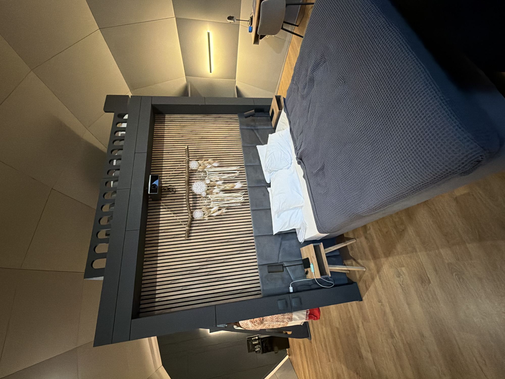 The image depicts a modern hotel room in Solov'evo, Russia, featuring a large bed with a dark gray duvet and white pillows, situated against a wooden slat headboard. Above the headboard, a dark gray structure with arched detailing is visible, possibly a decorative element or part of the room's architecture. Dreamcatchers hang from a wooden branch above the bed. To the right, a desk with a chair and a lamp are set against a textured wall with a horizontal light fixture. A bedside table with a phone and a lamp is next to the bed on the left. In the background, a person wearing a patterned garment and red shoes is partially visible near a black appliance. The room has wooden flooring and a unique, geometric ceiling.