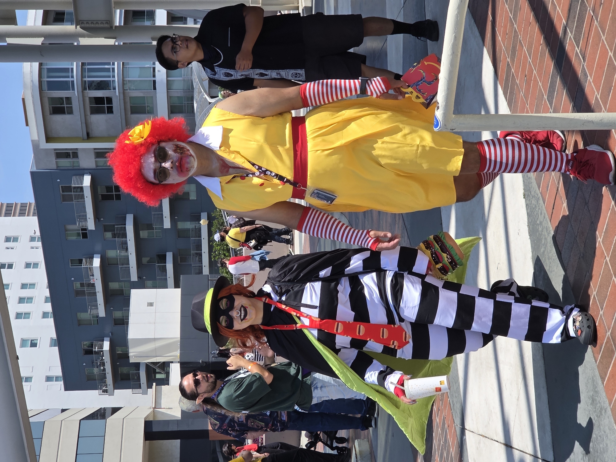 Two individuals in costumes are walking on a paved outdoor walkway in front of modern buildings. The person on the left is dressed as the Hamburglar, wearing a black and white striped jumpsuit, a black hat with a green band, a green cape, and a red tie with hamburger print. They are holding a white cup with a McDonald's logo. The person on the right is dressed as a zombie Ronald McDonald, wearing a yellow dress with a red belt, a red clown wig with a yellow flower, and red and white striped arm warmers and socks. They have zombie-like makeup with fake blood on their face and chest. A man in a black shirt and shorts, wearing glasses, walks behind them. The background shows buildings with balconies and windows, characteristic of urban architecture. The lighting suggests a bright, sunny day. The environment appears to be a public space in the city of Long Beach, California, United States of America, potentially at a convention or street fair.
