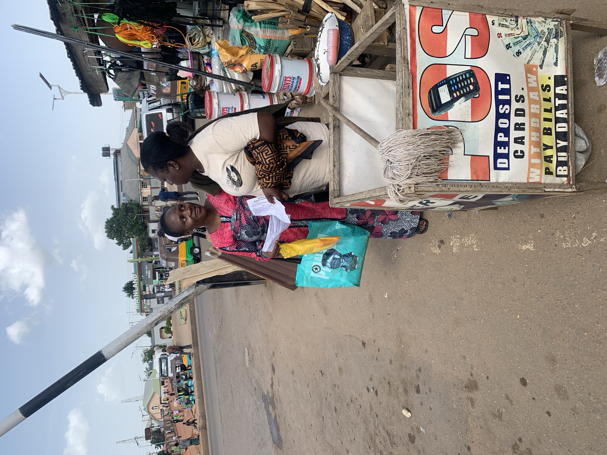 This image captures a street scene in Gura Top, Nigeria, likely during the daytime. Two women are the central subjects. One woman, wearing a light-colored shirt and a patterned skirt, is leaning over, possibly interacting with the other woman who is mostly obscured. The second woman is wearing a brightly colored, patterned dress.
The setting appears to be a busy marketplace or roadside stall. Various objects are visible, including stacks of paint buckets labeled "PAINTS EMULSION," bags of fertilizer marked "SPECIAL FERTILIZERS 50KG," rolls of rope in vibrant colors like orange and green, and what looks like construction materials. A prominent sign with red and white stripes features the word "Opay" and lists services such as "DEPOSIT CARDS," "WITHDRAW," "PAY BILLS," and "BUY DATA." There are also images of banknotes and a credit card terminal on the sign. A turquoise shopping bag with a cartoon character and a yellow plastic bag are placed on a surface in the foreground.
The ground is a dusty, paved surface with some stains. In the background, buildings and vehicles, including what appears to be a truck and a tricycle (Keke Napep), suggest a lively urban environment. The sky is partly cloudy, indicating typical weather conditions. The overall impression is a snapshot of everyday commerce and activity on a Nigerian street.