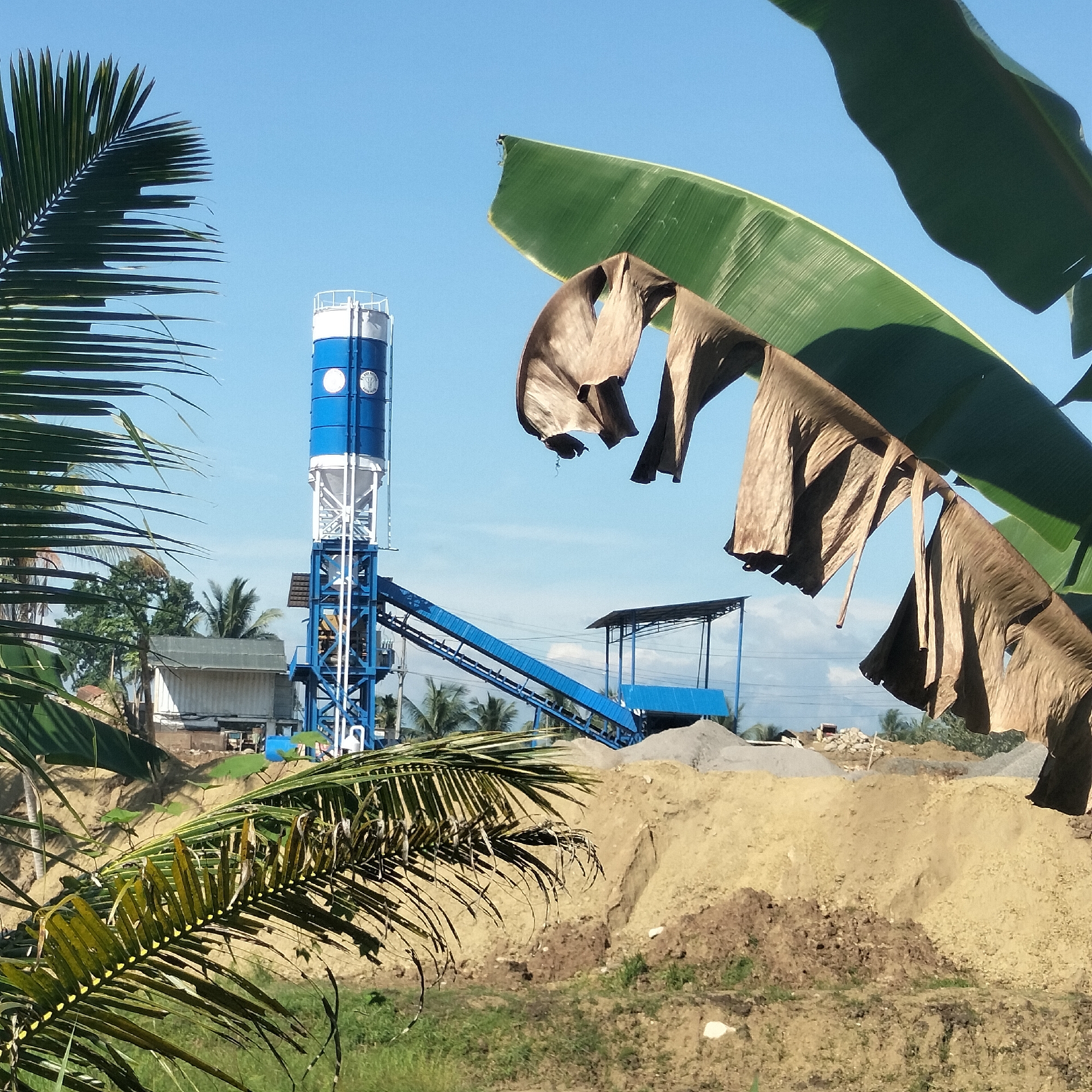 This media file captures an industrial scene, likely a concrete batching plant, under a bright, clear blue sky in Seuneubok, Indonesia. The central subject is a tall, blue and white cylindrical tower, characteristic of a cement silo, supported by a metal framework. A blue conveyor belt system extends diagonally from the lower part of this tower towards the right, leading to mounds of aggregate materials such as sand and gravel piled on the ground.
The foreground and sides of the image are framed by lush tropical vegetation. On the left and bottom, vibrant green palm fronds are visible, while large banana leaves, some green and others dried and brown, hang from the top and right, partially obscuring the view. In the background, other palm trees and a simple building with a light-colored roof can be seen.
The scene suggests an ongoing industrial operation for producing construction materials. The bright sunlight and absence of clouds indicate a clear daytime setting, likely mid-morning or afternoon, with strong natural lighting. A circular emblem or logo is visible on the blue section of the main cylindrical tower, though the text within it is not decipherable. No people are visible, and the scene is devoid of overt activity, presenting a static view of the industrial site within its natural surroundings.