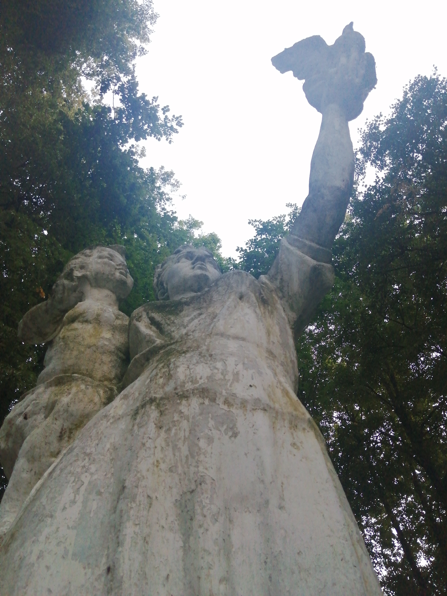 An upward-angle shot captures a stone sculpture of two figures, set against a backdrop of green foliage and an overcast sky. The larger figure, on the right, stands with its right arm raised, holding a bird or a similar object aloft. The figure on the left, appearing smaller and possibly a child, is positioned beside and slightly behind the larger figure, with its left arm extended. The sculpture is weathered, showing patches of moss and a rough texture. The scene is set in Slonim, Belarus, and is likely located in a park or public space.