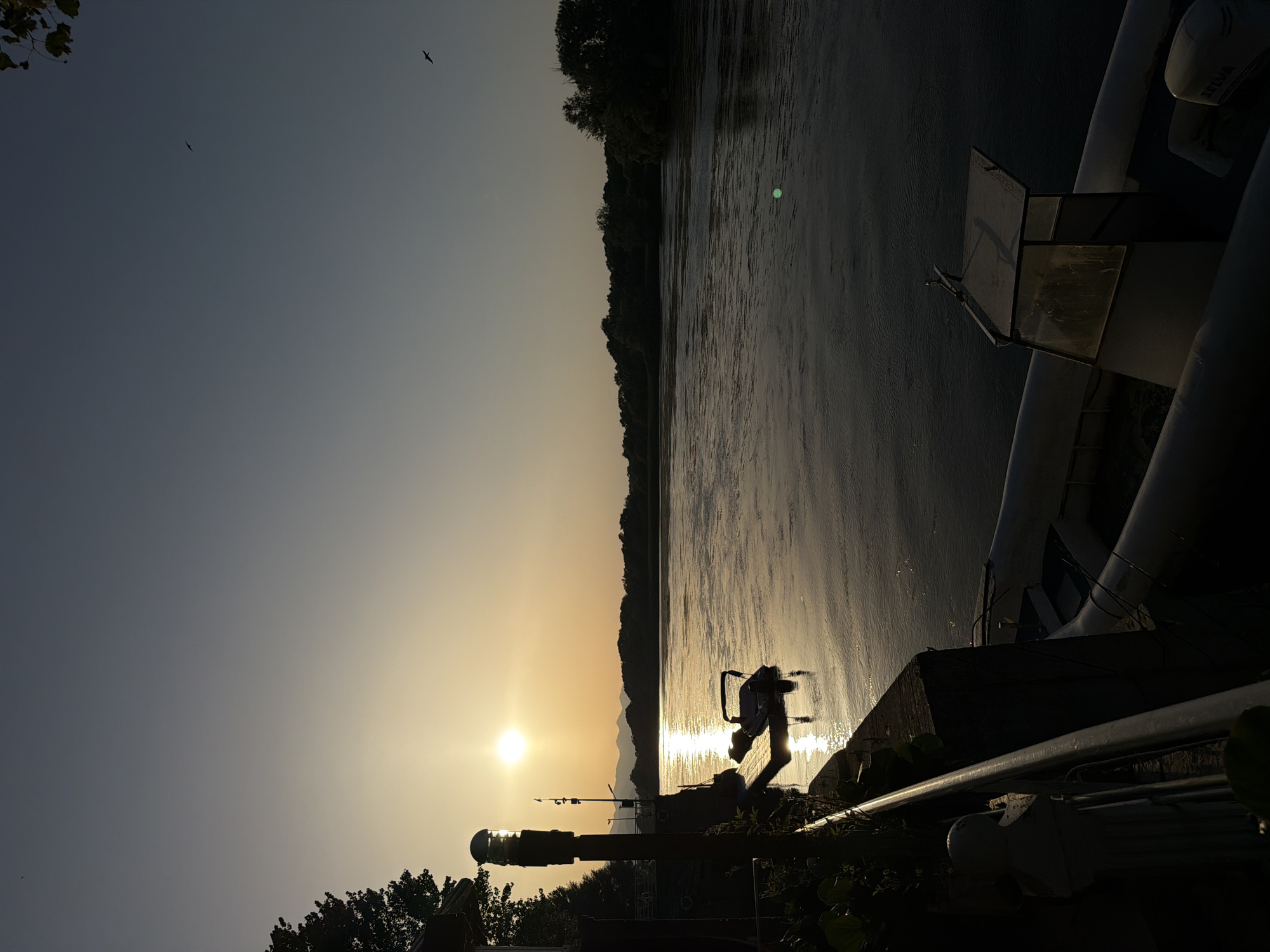 This is a vertical, medium shot of a river during sunset in Shkoder, Albania. The sun is low on the horizon, casting a warm glow across the water and sky. The water is calm, with ripples reflecting the sunlight. Trees and mountains form a dark silhouette in the background. In the foreground, a dock is visible with a small boat tied to it. A lamppost stands on the left side of the frame, illuminated by the setting sun. The scene is peaceful and serene, capturing the beauty of a sunset over a river.