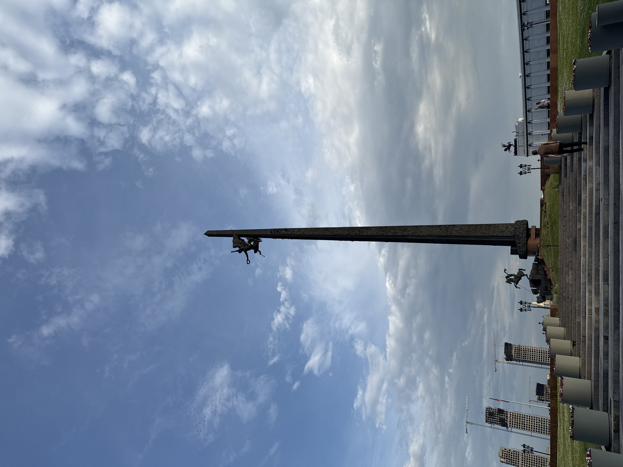 The image shows the Victory Monument, also known as the Monument to the Heroes of the Soviet Union in the Great Patriotic War, located in Victory Park on Poklonnaya Hill in Moscow, Russia. The monument is a tall, dark obelisk topped with a sculpture of Nike, the Greek goddess of victory, and two winged figures. At the base of the monument, there is a sculpture of a rider on horseback.
In the background, there are several modern buildings under construction, with cranes visible. In the middle ground, there are concrete planters filled with flowers, a wide set of stairs leading up to the monument, and lampposts. A few people are visible in the scene, including one person walking up the stairs and others standing on the grass in the distance. The sky is partly cloudy, with a mix of blue sky and white and gray clouds. The overall lighting suggests it is either early morning or late afternoon.