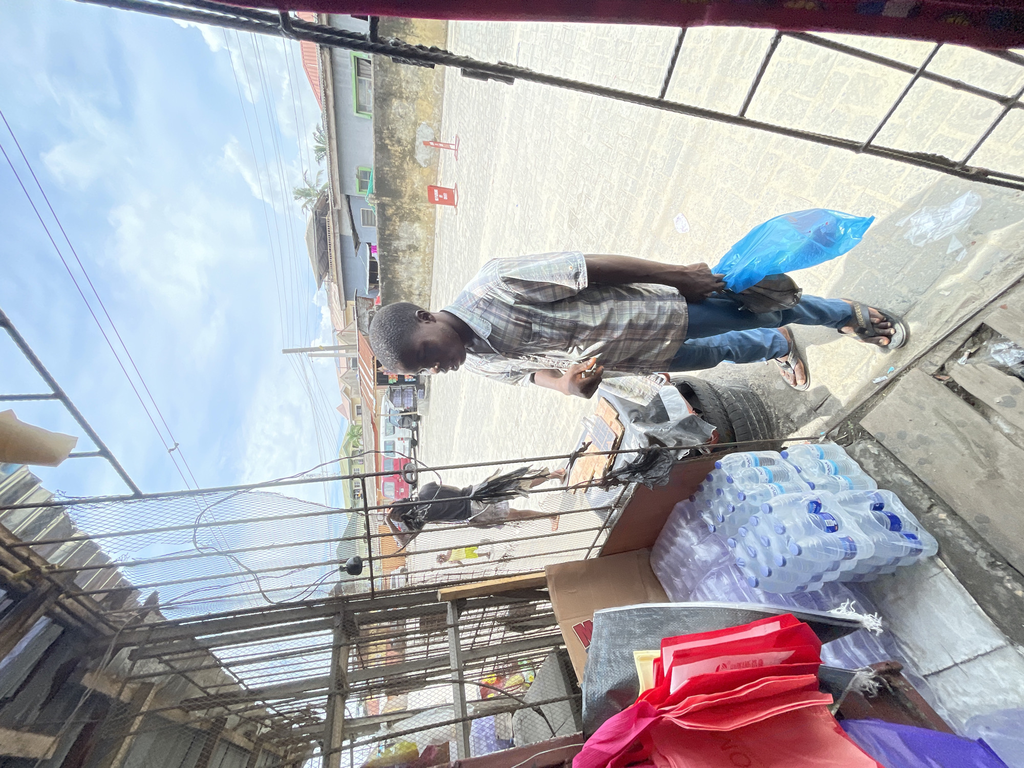 The image, taken in Ikorodu, Nigeria, shows a man wearing a plaid shirt and jeans, holding a blue plastic bag. He is standing on a paved surface, possibly a street or sidewalk. He is viewed from behind a caged enclosure, with a cluttered foreground containing stacked bottled water, shopping bags, cardboard boxes, and debris. The background includes buildings and vehicles on the street. The sky is partly cloudy.