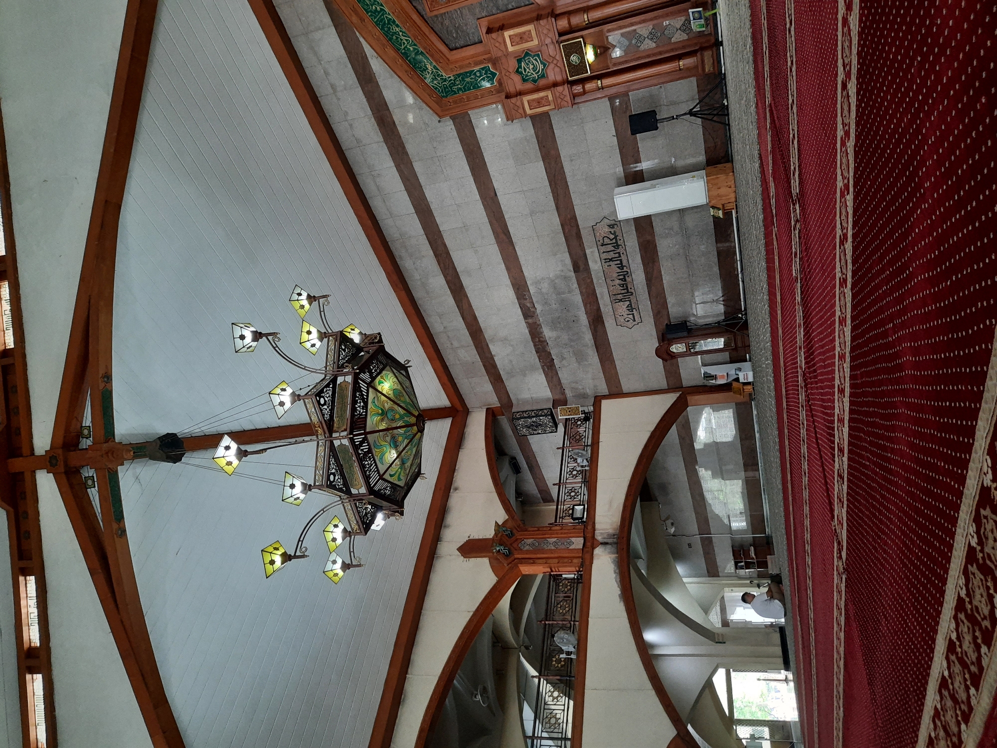This image depicts the interior of a mosque, likely located in Bandung City, Indonesia, with a focus on its architectural features and furnishings. A large, ornate chandelier with multiple lamps hangs from the high, wooden-beamed ceiling. The walls are paneled with alternating bands of grey stone and darker wood. The floor is covered with a richly patterned red carpet, indicative of prayer mats.
In the foreground, the red carpet is prominently featured, with its intricate design suggesting designated prayer spaces. In the mid-ground, a man in a white shirt is seated on the carpet, facing away from the camera, near an open doorway. Arching structures and balconies are visible in the background, suggesting a multi-level interior. Islamic calligraphy adorns the wall above a white air conditioning unit and a speaker on a tripod. To the right, a large wooden structure, possibly part of the mihrab or a decorative element, is visible with green Islamic motifs. The overall impression is one of a serene and well-maintained place of worship.