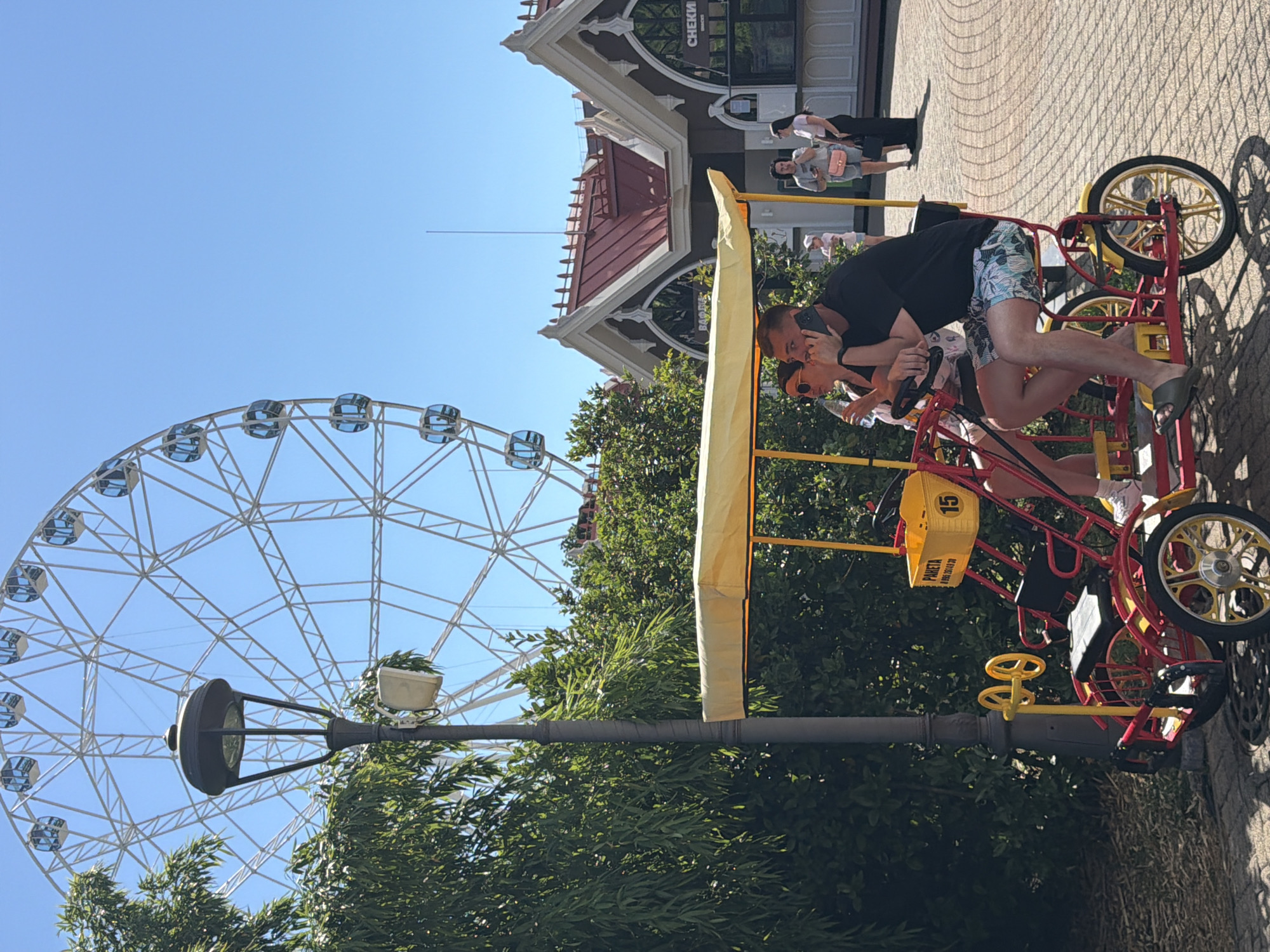 In Sochi, Russia, under a clear blue sky, a couple is enjoying a sunny day at an amusement park. They are seated on a bright red quadricycle, which appears to be a pedal-powered vehicle designed for multiple passengers. The man, wearing a black t-shirt and patterned shorts, is looking at his phone while holding onto the steering wheel. The woman, seated next to him, leans in to kiss him. Her attire is not clearly visible.
The quadricycle has a yellow canopy, offering shade from the sun. A yellow basket is attached to the back, with the number "15" visible on it and some text that reads "PARETA" and a phone number. The vehicle's wheels are yellow and silver.
In the background, a large Ferris wheel with numerous cabins is prominent against the sky. Adjacent to it is a building with a distinctive architectural style, featuring a red-tiled roof and dark decorative trim, suggesting it might be part of the amusement park's infrastructure. Several other people are walking around in the background, some of whom are casually dressed, indicating a leisurely atmosphere. A lamppost is positioned in the foreground, partially obscuring the view of the Ferris wheel. Lush green trees provide a natural element to the scene. The overall mood appears to be one of joy and relaxation, characteristic of a day out at an amusement park on a pleasant day.
