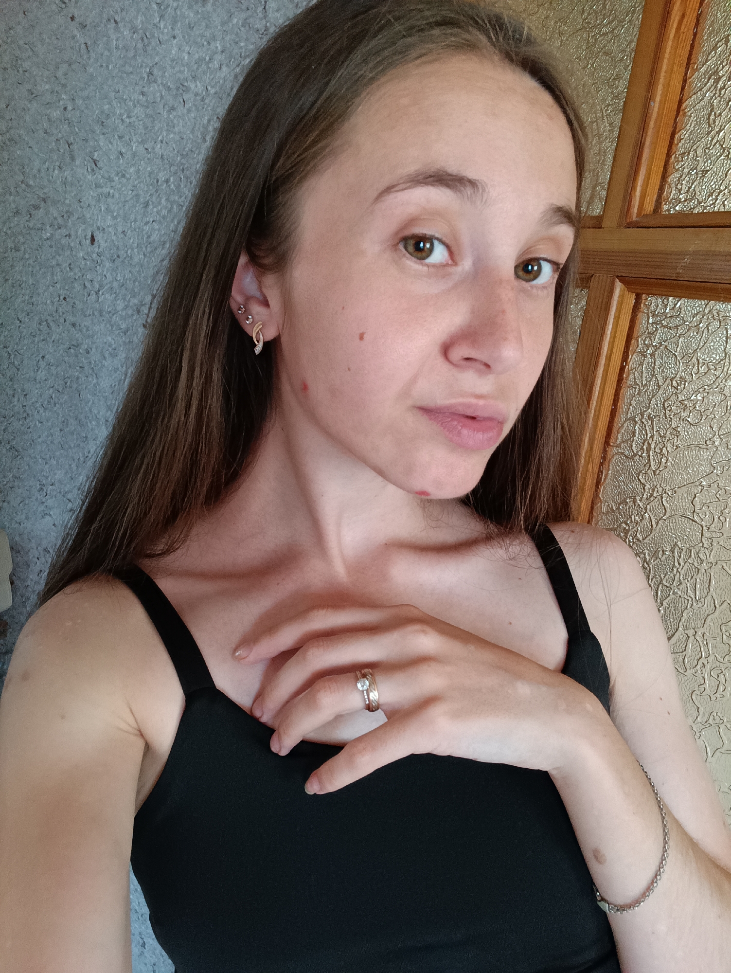 A young woman with long, dark brown hair is photographed from the chest up, facing slightly to her right. She is wearing a black tank top. Her left hand rests on her chest, and she is wearing a ring on her right index finger. She has several piercings in her left ear. A textured gray wall is visible behind her to the left, and a wooden door with textured glass is to her right. The image appears to be taken indoors, possibly in a residential setting in Vypasne, Ukraine.