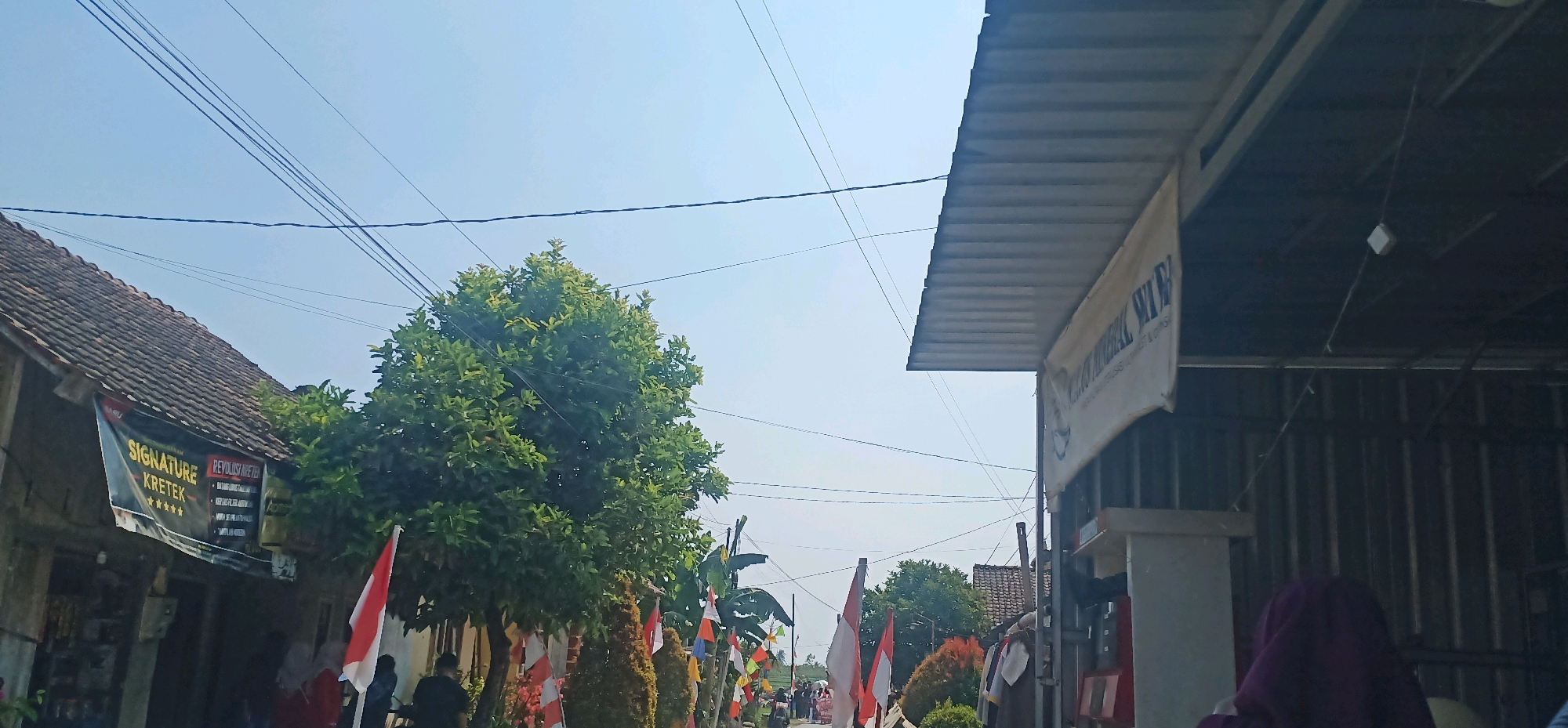 The image depicts a street scene in the city of Kincang, Indonesia. A building with a metal roof is on the right, displaying a white banner with text. A large green tree stands in the middle, surrounded by various small decorative plants. Several Indonesian flags, red and white, are displayed along the street, suggesting a celebratory or patriotic event. Several people are visible in the background, some appearing to be walking or standing. A banner on a building to the left advertises "SIGNATURE KRETEK". The sky is clear and blue. The environment suggests a residential or commercial street in a tropical climate.