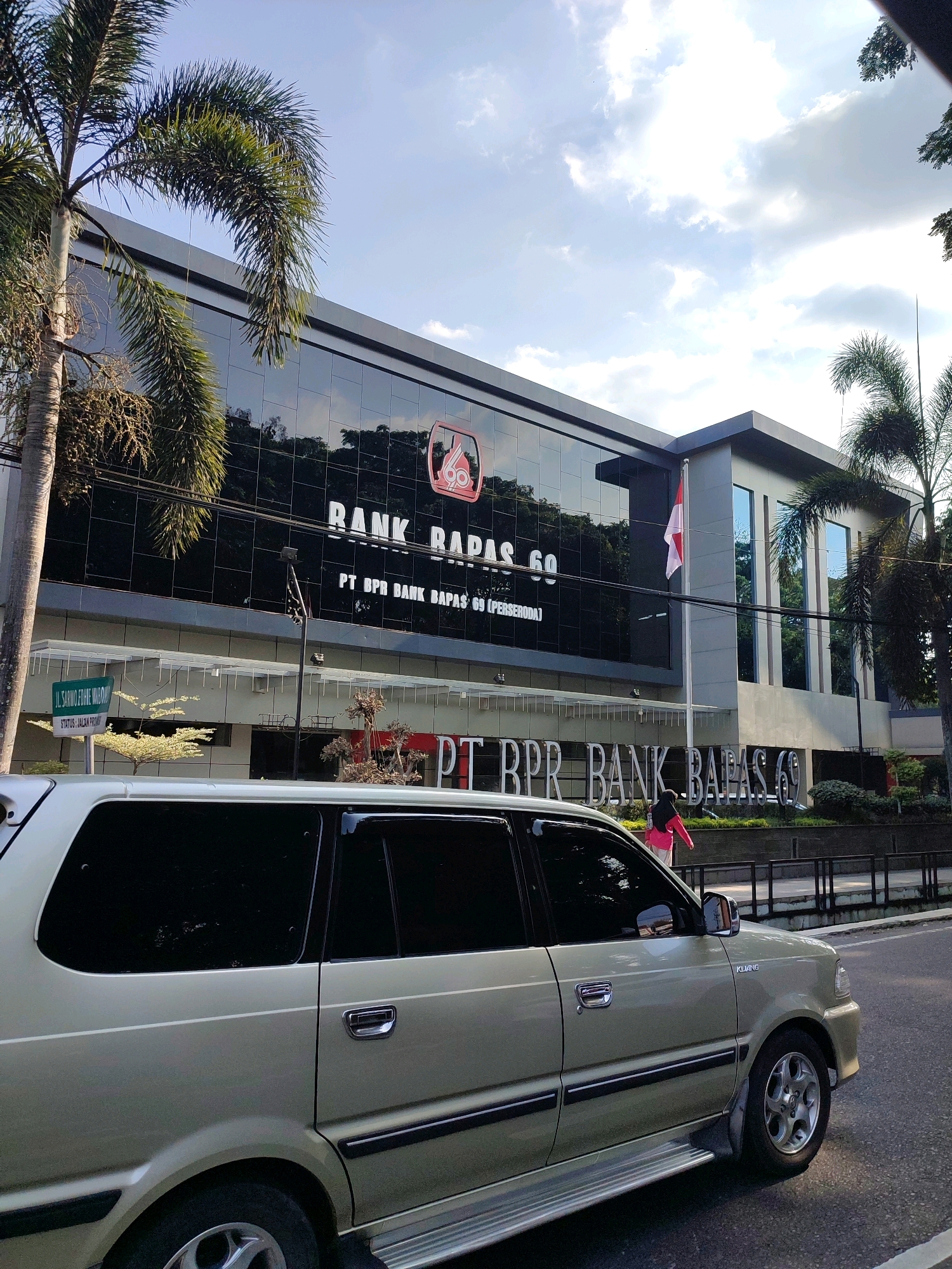 A light-colored minivan is parked on a street in front of a modern building with a glass facade. The building has the text "BANK RAPAS 69" and "PT BPR BANK RAPAS 69 (PERSERO)" on its exterior. An Indonesian flag is flying on a flagpole in front of the building. Palm trees are visible on the left and right sides of the frame, framing the building and the street. The sky is partly cloudy, with the sun casting shadows. A person in a pink jacket is walking in front of the building. A street sign is visible on the left, indicating "JL. SARNO FOHE WIDODO" and "STATUS: JALAN PROMO". The scene appears to be a street in the city of Banyurojo, Indonesia.