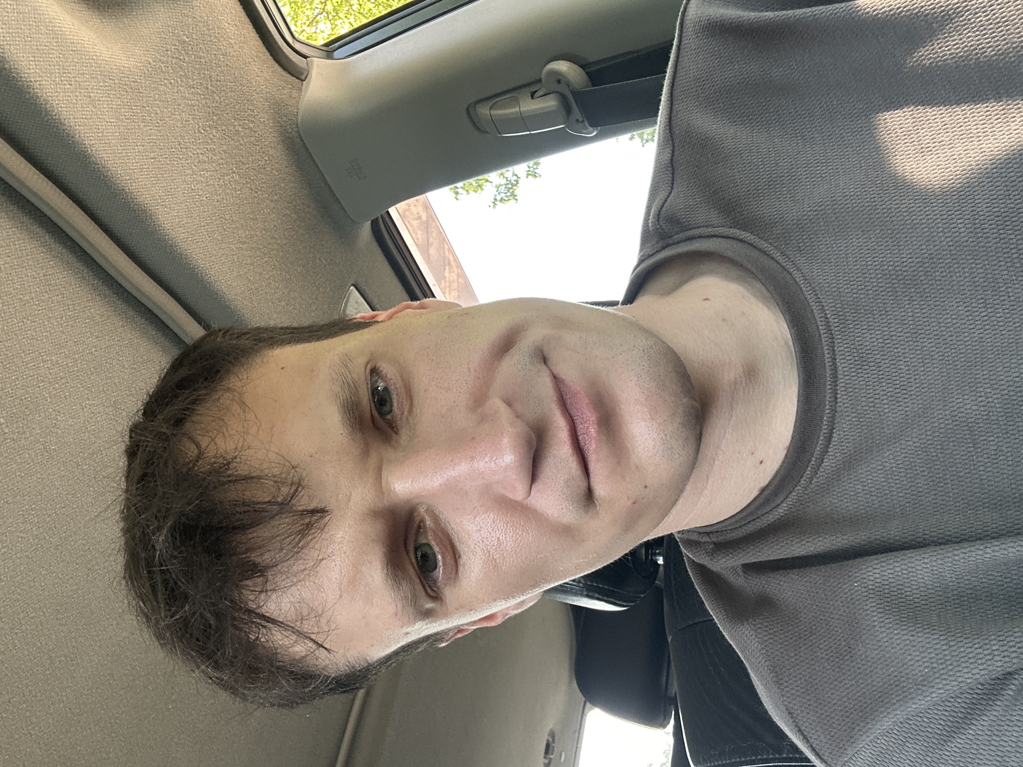 A close-up selfie, taken from a low angle, shows a person sitting in the passenger seat of a vehicle, with the headrest of the driver's seat visible behind. The person has light skin, short, dark brown hair, and is wearing a gray, textured t-shirt. Sunlight illuminates the right side of their face and shoulder, creating a shadow on their shirt. The background reveals the car's interior, including the ceiling, a seatbelt, and a glimpse of green foliage through the window, suggesting an outdoor setting. The image does not contain any discernible information that would indicate its location in the city of Makiv, Ukraine.