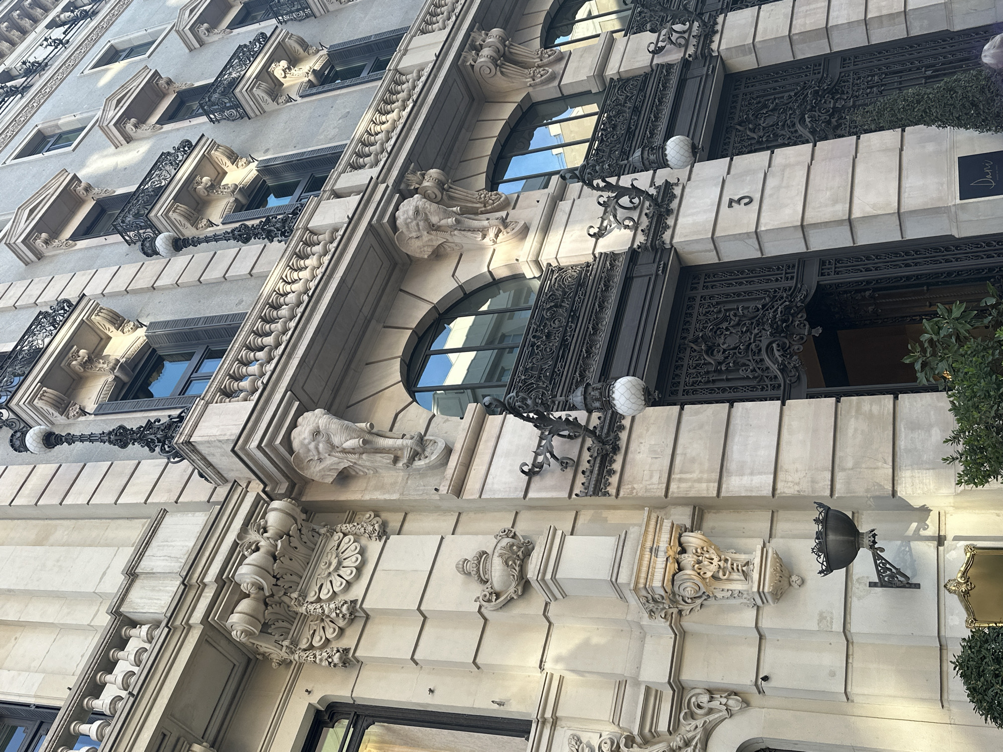 The image shows the exterior facade of a building in Madrid, Spain, taken from a low-angle perspective. The architecture is characterized by ornate detailing, including elephant head sculptures, decorative ironwork balconies, and elaborate molding. The building has a light-colored stone facade, with the number "3" visible above an entranceway. A sign with the name "Dani" is also present. Street lamps with decorative ironwork are attached to the building. The windows are arched, reflecting the sky.
