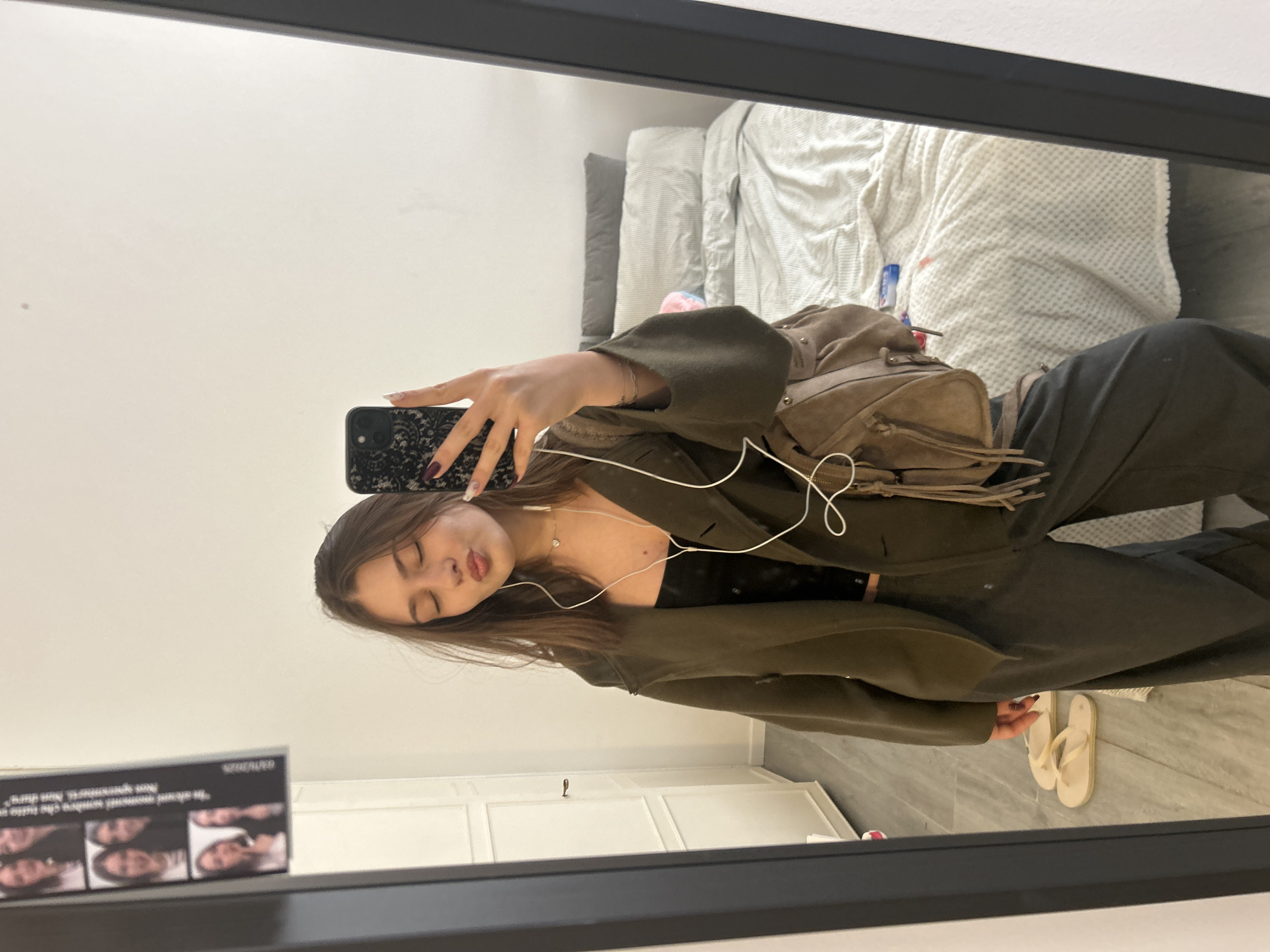 A young woman is taking a mirror selfie in what appears to be an apartment. She is wearing an olive green blazer over a black top, and dark trousers. She has a brown backpack slung over her shoulder. She is holding a black smartphone, and she has white earphones plugged into her ears. Her eyes are closed and she has slightly pursed lips.
In the background, there is a white wall, a bed with a patterned white duvet, and white cupboards. There are also a pair of flip flops on the floor.
The location cannot be determined from the image, but I will assume she's in the city of Milan, Italy.