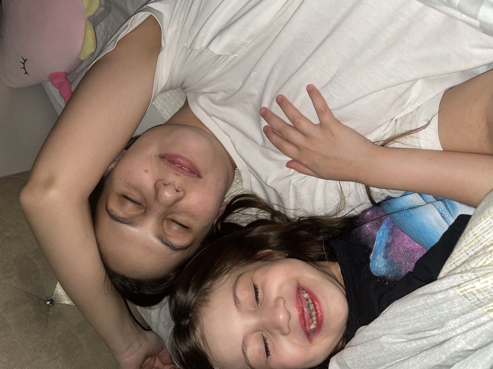 In a dimly lit domestic bedroom in Izhevsk, Russia, an adult female and a young female child are depicted lying side-by-side on a bed. Both individuals have their eyes closed.
The adult female, positioned on the right side of the frame, is lying on her back with her head oriented towards the right. Her dark hair is spread beneath her head. She exhibits a subtle smile with her eyes closed. Freckles are visible across her nose and cheeks. She is wearing a white short-sleeved t-shirt. Her right arm is bent, with her forearm and hand positioned above her head, resting against a beige, textured headboard featuring a single decorative crystal-like stud.
The child, positioned on the left side of the frame, is lying on her back with her head oriented towards the left. Her long brown hair frames her face. She has a wide, open-mouthed smile, revealing teeth with visible dental work (fillings or caries) and a dental appliance (possibly a retainer or braces) on her lower teeth. Her lips are a distinct red hue. She wears a black long-sleeved shirt featuring a multi-colored graphic design, possibly depicting a galaxy or cosmic scene. Her left hand is resting on the adult's chest area.
The individuals are covered by light-colored bedding, appearing to be sheets or a duvet. Behind the adult's left shoulder, a pink unicorn plush toy with a pink bow and yellow feet is visible on the bed. The overall lighting is soft and diffused, indicative of an indoor, low-light setting.