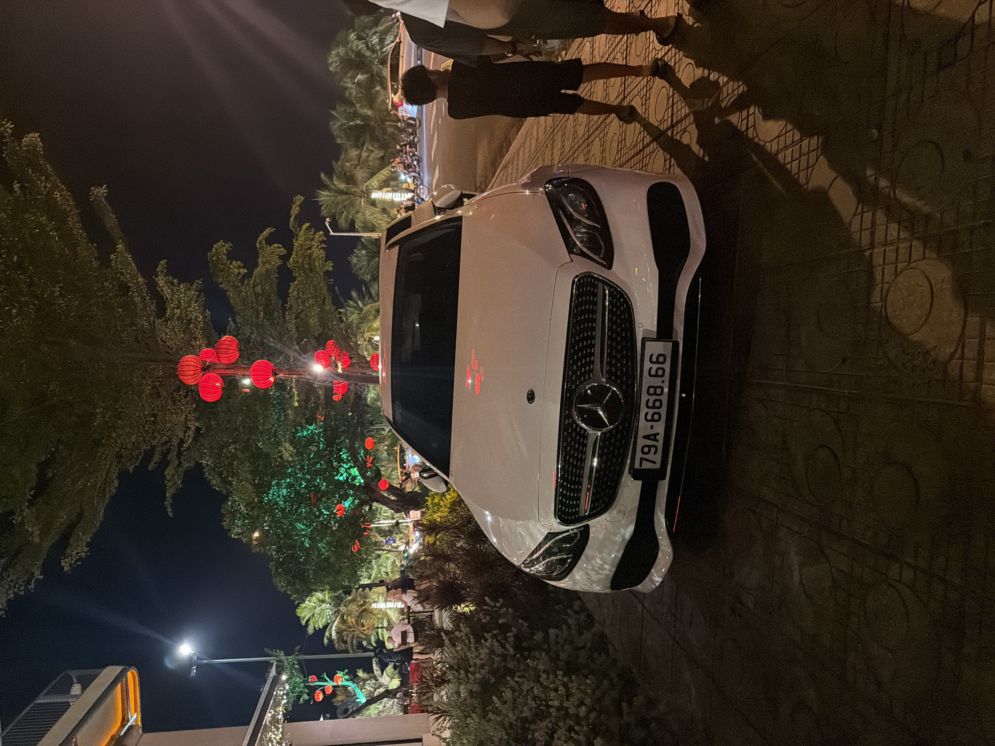 This detailed description of the image is based on forensic, matter-of-fact observations:
The image depicts a nighttime urban street scene in Nha Trang Ward, Khánh Hòa Province, Vietnam. In the foreground, a white Mercedes-Benz sedan with Vietnamese license plate "79A-668.66" is parked on a patterned concrete sidewalk, facing the camera. Its front grille and headlamps are clearly visible.
To the right of the parked vehicle, two individuals, an adult and a child, are partially visible, seen from behind. The child, dressed in a dark t-shirt and shorts, stands to the left of the adult, both appearing to face towards the street or right side of the frame. Their shadows are cast prominently on the sidewalk directly in front of them, extending towards the lower right.
The environment is characterized by a combination of natural and artificial elements under dark skies. Numerous trees, including palm trees, line the street and are adorned with red spherical lanterns, providing decorative illumination. Some trees also have green uplighting on their foliage. A tall streetlight with a bright white lamp is visible on the left side of the frame, behind a multi-story building structure. This building, partially visible on the far left, exhibits warm strip lighting along what appears to be an upper floor or balcony. Below this, an outdoor patio or dining area is discernible, with several indistinct figures seated or standing.
In the mid-ground and background, beyond the Mercedes and people, a paved road is visible with several vehicles, including motorbikes, and other pedestrians. The general ambiance is one of an active urban area at night, characterized by artificial lighting and tropical vegetation.