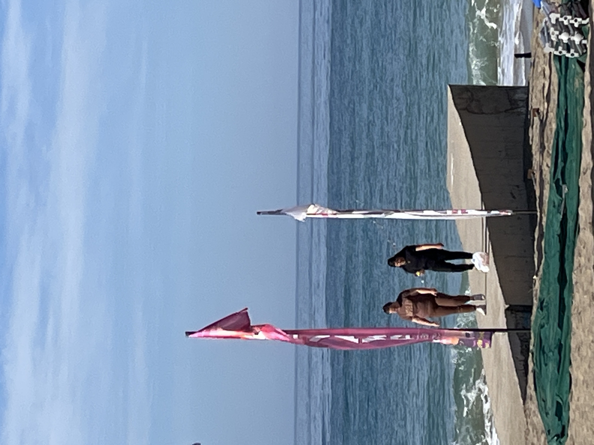 The image depicts a sunny day at a beach in Alanya, Turkey. The scene is oriented vertically with the sky dominating the left side, transitioning into the blue-green sea. Two people are standing on a concrete pier or walkway adjacent to the beach. One individual is dressed in brown clothing, while the other is in black. Pink and white banners are fixed in the sand/sidewalk, along the side of the frame on poles. The sea shows slight wave action, and the pier leads to a sandy area and a green net-like structure on the right edge of the image.