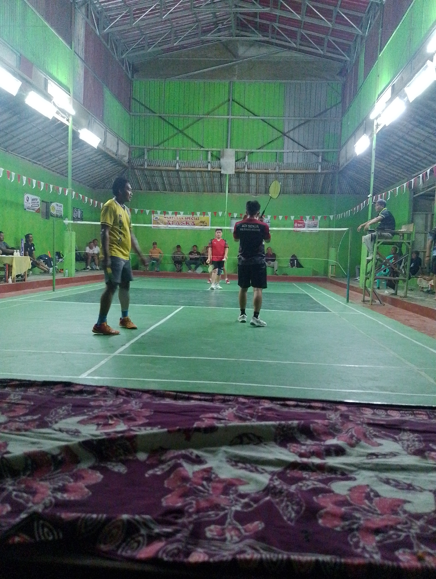 A badminton match is underway in a covered court at Adiraja, Indonesia. Two male players are on the court, one in a yellow shirt and shorts, the other in a black shirt and shorts. The player in the yellow shirt is looking towards the other side of the court, while the player in the black shirt is facing away, holding a racket. Several spectators are seated along the side of the court, and a person is seated in a raised chair, seemingly officiating. The court is green with white lines, and a net divides the two sides. The surrounding walls are painted green, and the roof is made of corrugated metal and other materials, supported by a metal frame. Fluorescent lights are suspended from the ceiling. In the foreground, a patterned cloth is visible.