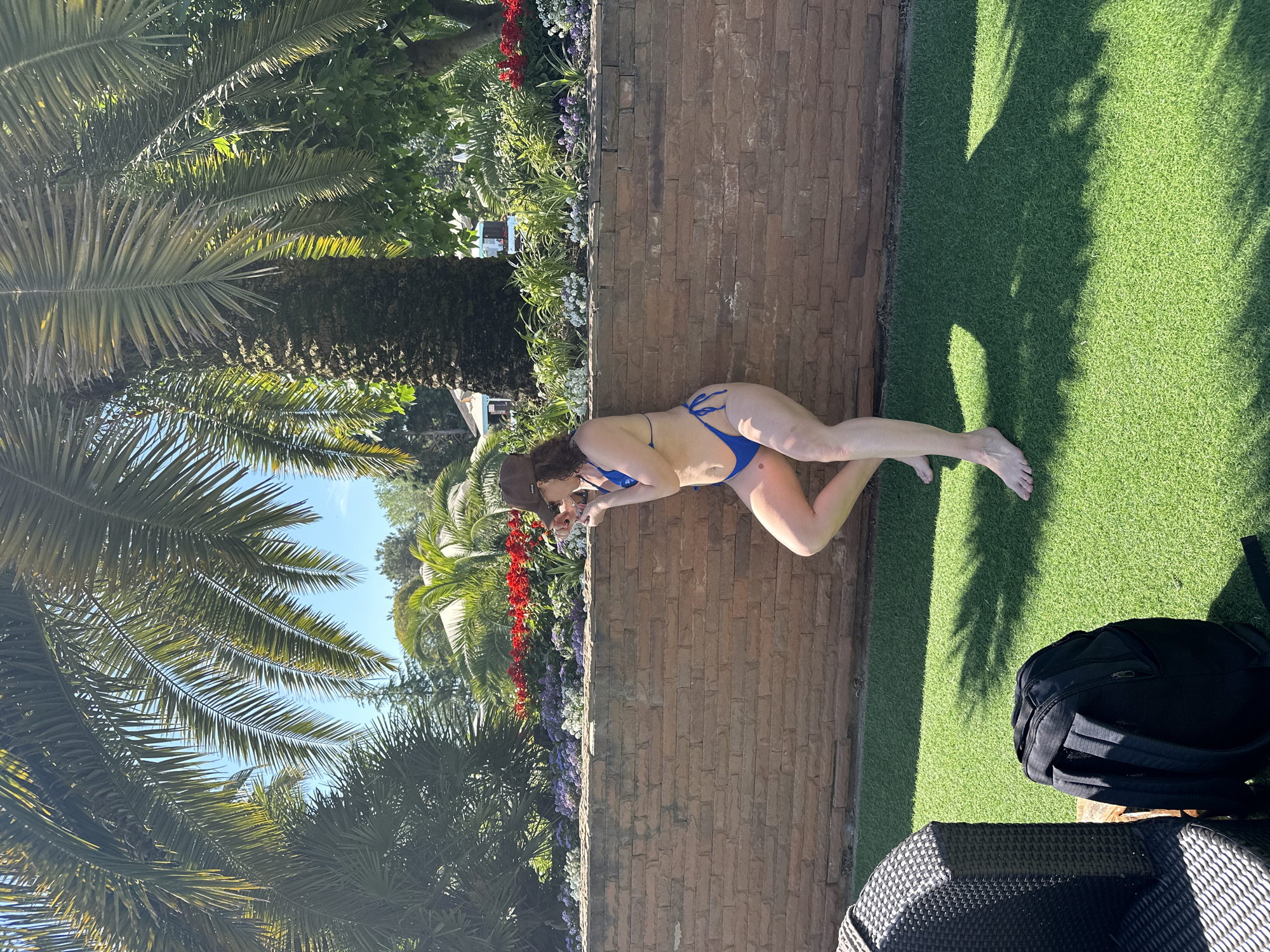 A middle-aged woman wearing a blue bikini and a brown baseball cap is lying on a brick wall. She appears to be looking down at something on the other side of the wall. To her right is a patch of artificial green grass and a black backpack. In the background, there are palm trees, a brick wall, red and purple flowers and blue sky. The photo was taken in the city of Sochi, Russia.