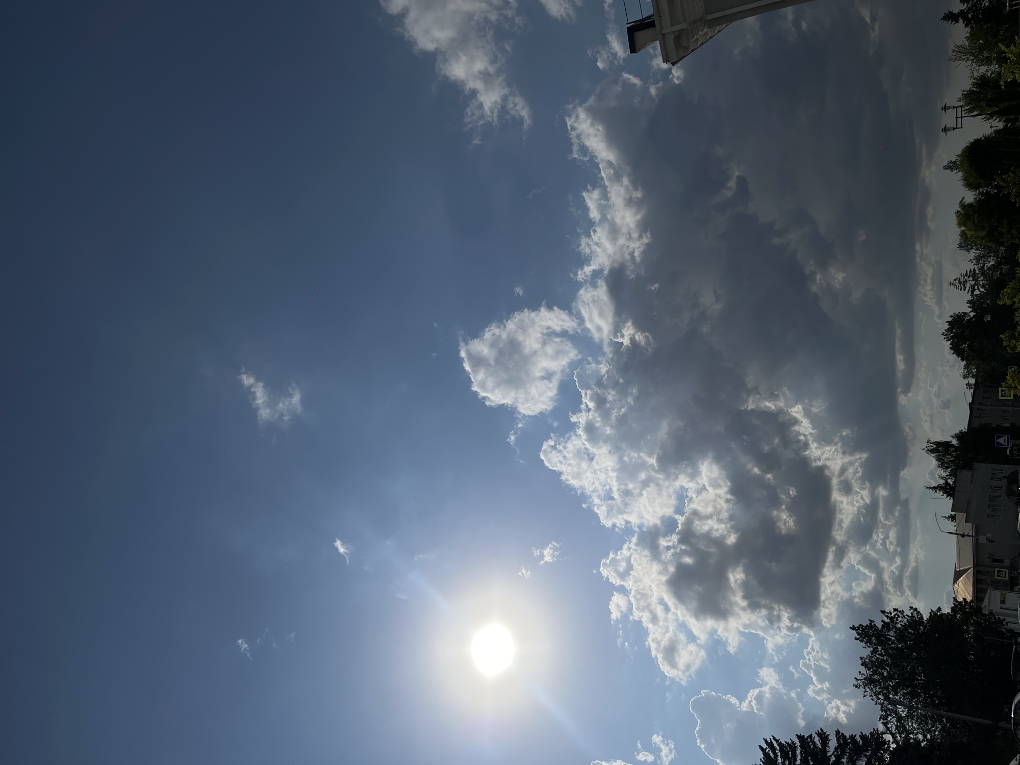 The image depicts a partly cloudy sky above a city scene in Novopokrovskaya, Russia. The sun is bright and positioned in the upper left quadrant, casting a glare and illuminating some of the clouds. The clouds are varied, with some appearing bright white and fluffy, while others are darker and more voluminous, suggesting potential for rain. In the lower portion of the image, the tops of buildings and trees are visible, indicating an urban or semi-urban environment. A few streetlights and traffic signs can also be discerned. The overall impression is of a bright, sunny day with dramatic cloud formations.