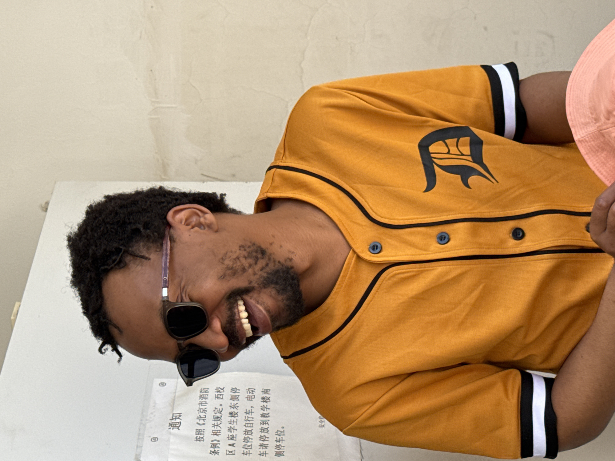 The image captures a man with dark, curly hair and a beard, wearing dark sunglasses and a bright yellow button-up shirt with black trim and a black "D" logo, resembling a baseball jersey. He is smiling widely, showing his teeth, and appears to be laughing or posing cheerfully. The man's head is tilted significantly, indicating the image is rotated. Part of another person wearing a pink hat is visible on the far right of the frame, suggesting a shared moment or interaction.
The setting appears to be outdoors or in a brightly lit indoor space, against a light-colored, textured wall. To the man's left, a white notice board displays Chinese text. The visible text, labeled "通知" (Notice), details regulations: "According to the relevant regulations of the 'Beijing Fire Control Regulations'. Bicycles parked in the parking spaces on the east side of Building A of the West Campus student dormitory, electric vehicles should be parked in the parking spaces on the south side of the teaching building." A partial phrase, "安全产" (Safety Production), is also discernible.
The presence of the notice board with Chinese text, specifically mentioning "Beijing Fire Control Regulations" and a "West Campus student dormitory" within a teaching building context, strongly suggests the location is a school or university campus in China, specifically in the Daxing District as per the provided context. The time of day appears to be daytime, with ample light. The man's joyful expression conveys a lighthearted and positive emotion.