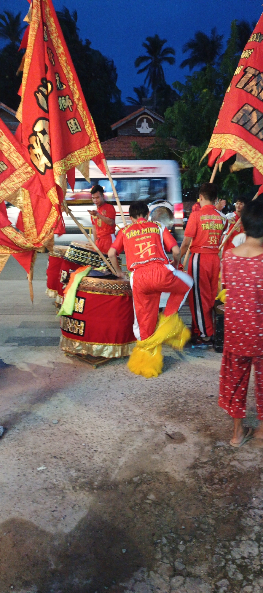 The scene captures an outdoor performance or gathering at dusk or night in the city of Unknown, Unknown. Several individuals, primarily men, are visible, many dressed in red uniforms.
In the center-left, a male individual wearing a red short-sleeved shirt and matching pants with white side stripes is positioned behind a large, cylindrical red drum adorned with gold trim and a black patterned band. The back of his shirt prominently displays the text "TAM MINH" in white capital letters above a stylized white character. He appears to be actively engaging with the drum, with a yellow, feathery object—likely part of a lion dance costume—near his left foot. To his immediate right, another male individual in a similar red uniform with black side stripes, also bearing "TAM MINH" on his back, stands facing away from the camera. Further left and partially obscured, a third male in red attire stands by another drum, appearing to hold an object to his mouth. On the far right foreground, a person, possibly a child, wears red and white patterned pajamas or loungewear, with their bare feet in light-colored flip-flops. In the mid-background, another individual in a red uniform is partially visible, smiling.
Three large, red triangular flags with ornate gold borders and black or gold characters are prominent. One on the far left displays vertical characters reading "藝聯醒獅" (Yì Lián Xǐng Shī, commonly translated as "Art Unite Waking Lion" or "Art Association Lion Dance"). Another flag, partially visible on the right, appears to have "TAM MINH" text. The drums are red with gold accents and some visible text, including "MINH".
In the background, a white bus with blurred red text passes horizontally, indicating motion and a street environment. Behind the bus and flags, a building with a dark tiled roof is visible. Its gable features a distinct white emblem depicting a figure in a meditative pose, likely a Buddha or Bodhisattva. Dark foliage and palm trees surround the building and extend against a dark blue evening sky. The ground is a rough, uneven surface of concrete or asphalt, showing varying shades of grey and brown, with some darker, wet-looking patches. The scene is illuminated by artificial light sources not visible in the frame, casting some shadows and highlighting the vibrant red colors of the costumes and flags.