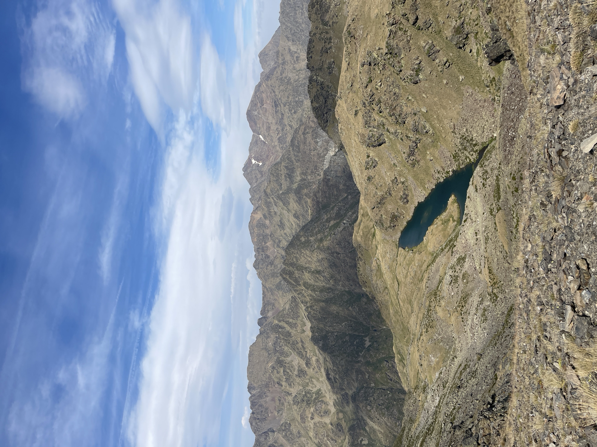 An eye-level, high-angle medium outdoor shot captures a mountainous landscape under a blue sky with scattered white clouds. In the foreground, a rocky, grassy slope descends, with scattered rocks and dry grass. Further down, a dark blue lake is visible, nestled in a valley. The midground and background are dominated by rugged, brown and green mountains, characterized by their rocky terrain and sparse vegetation. Some of the distant peaks show hints of snow. The lighting suggests it is daytime.