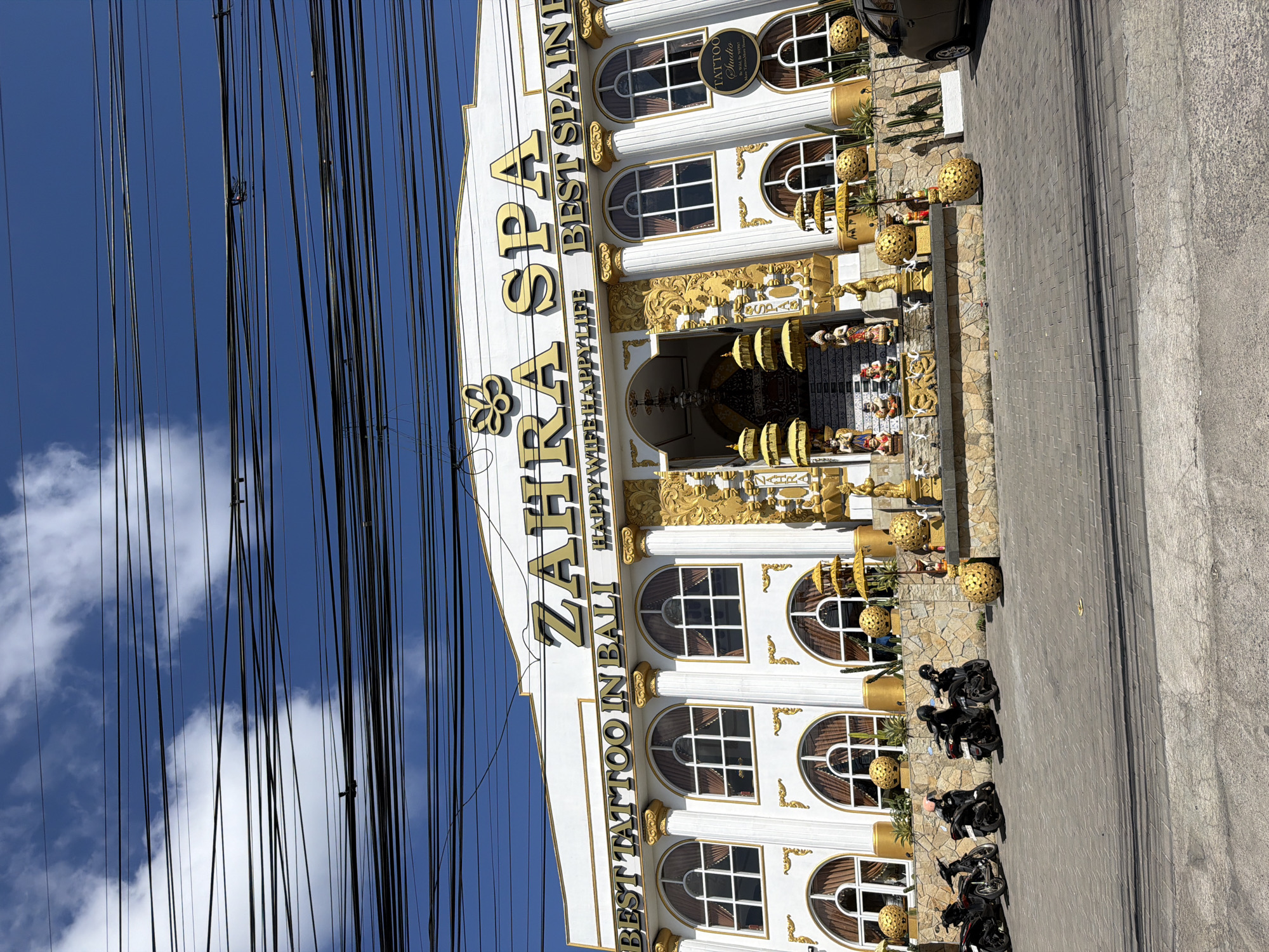 The image shows the Zahra Spa and Tattoo Studio located in Kuta Selatan, Indonesia. The building has a white facade with ornate gold decorations, including the spa's name in large gold letters and decorative columns. Multiple arched windows line the upper level, each with brown curtains. Below the windows, a stone wall serves as a base for the building, adorned with various gold statues and decorative elements, including what appear to be Balinese parasols. In front of the spa, there is a paved area with several motorcycles parked. To the right of the building, a dark-colored car is parked. The sky above is blue with white clouds, and a dense network of black power lines crisscrosses the upper portion of the image, obscuring parts of the sky and the top of the building. The overall scene depicts a commercial establishment on a street.