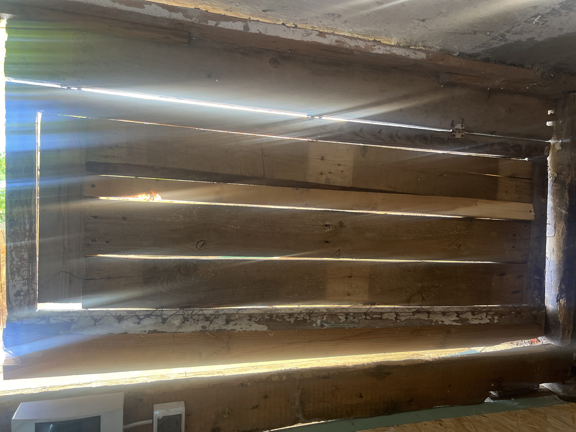 An eye-level, medium shot captures a rustic, wooden door with vertical planks, situated in what appears to be an interior space. Sunlight streams through gaps between the planks and from the top of the door, creating dramatic shafts of light that illuminate dust particles. The door is slightly ajar, revealing hints of a dimly lit area beyond. The construction of the door and its surroundings suggests an unfinished or dilapidated setting, with visible nails, peeling paint, and rough-hewn wood. A simple metal latch is attached to the door. The overall impression is one of aged, functional simplicity, with the intense sunlight adding a sense of drama to the scene.