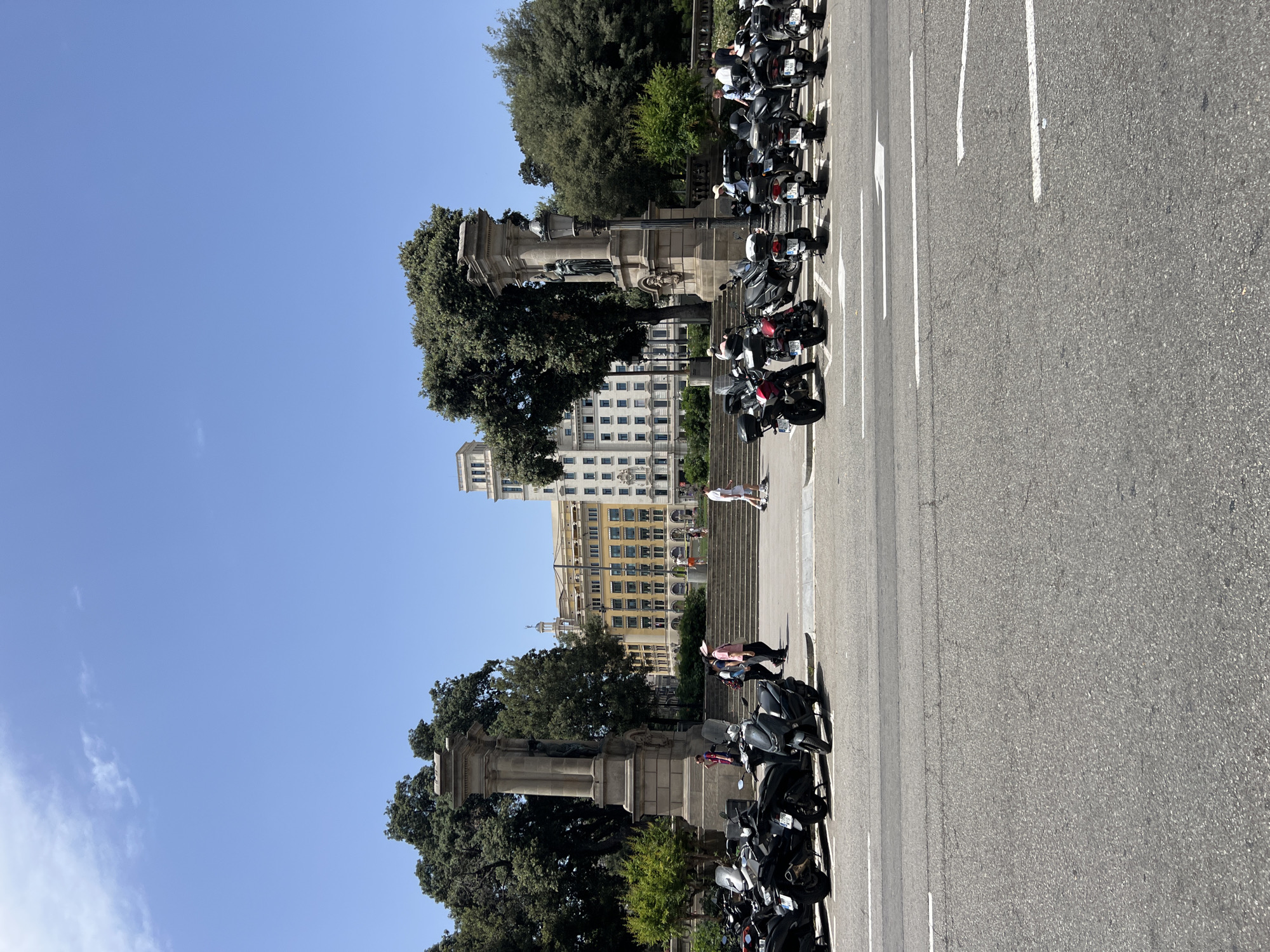 The image shows a street scene in Barcelona, Spain, on a clear, sunny day. The perspective is from above, looking down on a row of parked motorcycles. In the background, there is a large, ornate monument with trees and classical architecture. Further back, a multi-story building with many windows stands under a bright blue sky with a few wispy clouds. Several people are visible on the steps leading up to the building and walking along the sidewalk. The motorcycles are parked in a designated area on the side of the road, with white lines marking the parking spaces. The scene appears to be from the late morning or early afternoon, given the strong sunlight and shadows. There is no visible text in the image.