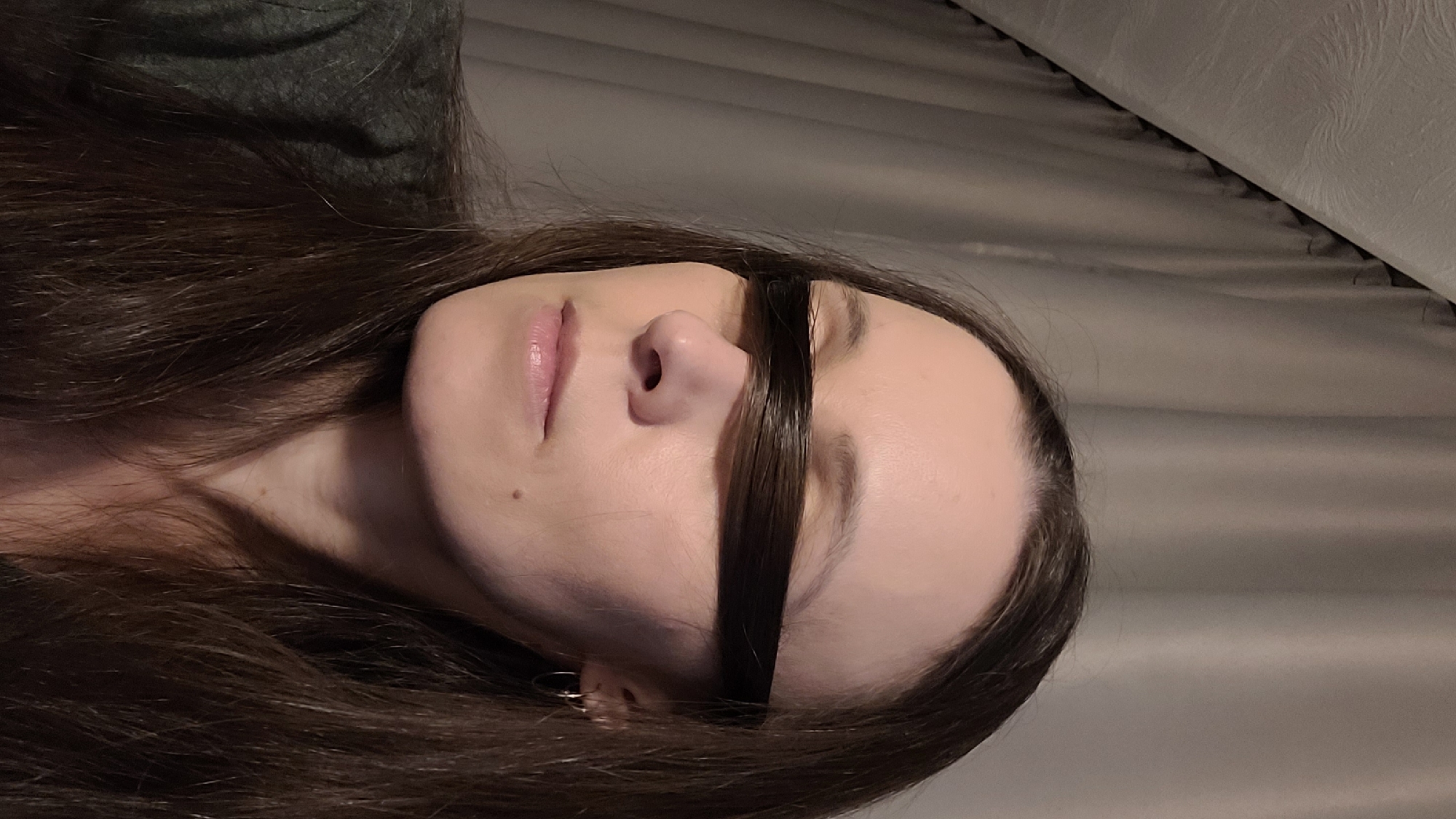 A medium shot captures a woman from the chest up, her eyes obscured by a thick band of her dark brown hair. She is positioned in front of a backdrop of draped, gray curtains. The woman is wearing a dark green, long-sleeved shirt. Her face is turned slightly to the left, with her eyes closed. A small mole is visible on her right cheek, and she has a small hoop earring in her right ear. The lighting is soft and appears to be coming from the front.