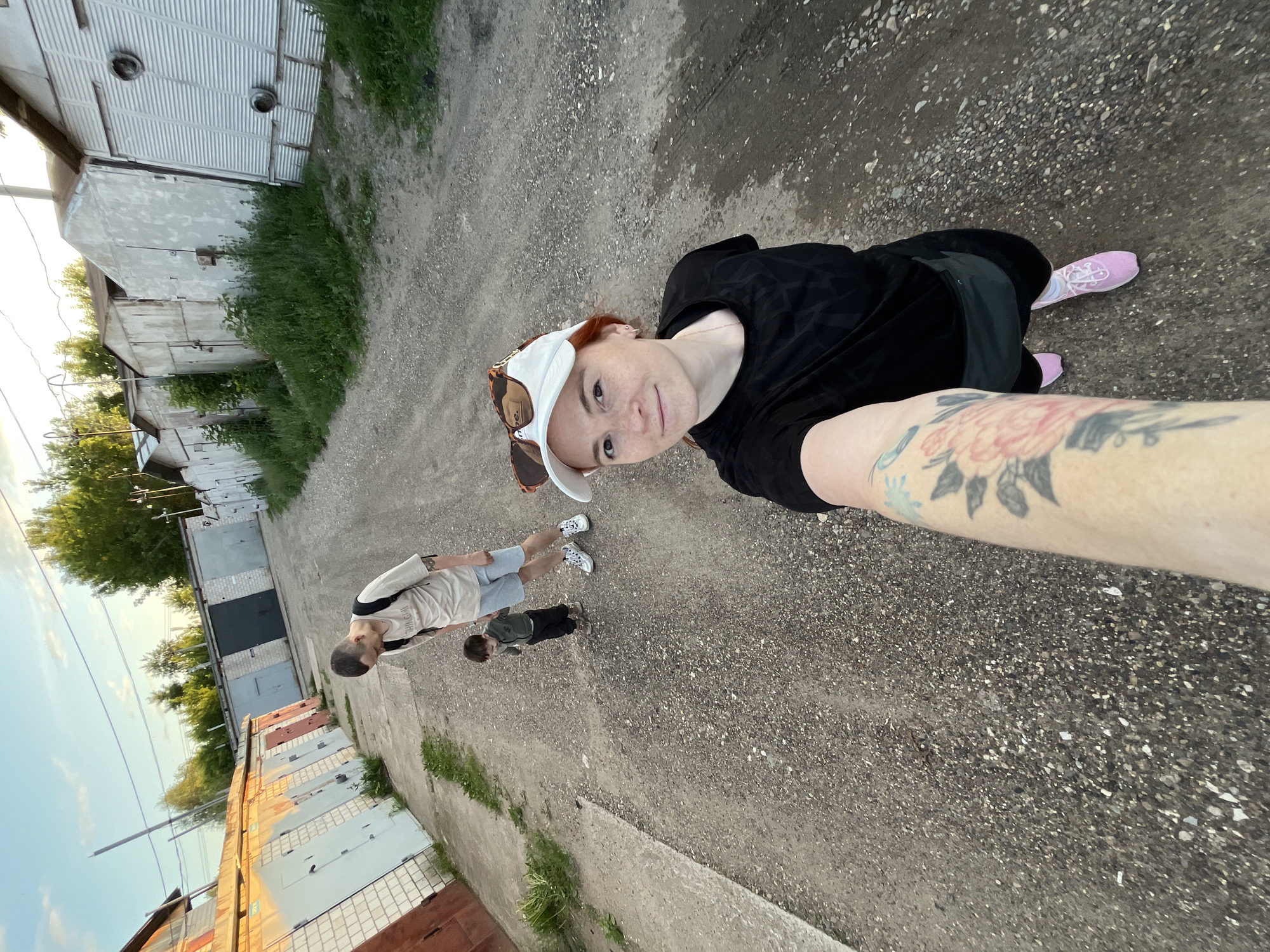 The image depicts a selfie taken by a woman on a gravel and dirt road in Chaikovsky, Russia. The woman, positioned in the immediate foreground, has reddish-brown hair, freckles, and wears a white baseball cap with tortoiseshell-framed sunglasses resting on top. She is dressed in a black short-sleeved top and dark lower garments, with visible tattoos on both forearms, including a prominent pink floral design on her right forearm. Her left arm, extended to hold the camera, also shows a blue tattoo. She wears pink sneakers.
Behind the woman, a man and a small child are walking on the same road. The man, with short dark hair and a slender build, wears a light beige t-shirt with dark lettering, light blue shorts, and white sneakers with black laces. He carries a dark backpack and is looking downwards towards the child. The child, a toddler, walks independently between the woman and the man, wearing a dark green long-sleeved top, dark pants, and brown shoes.
The environment consists of two parallel rows of single-story garages lining the unpaved road. On the left, the garages are varied in color (light blue, red, grey) and material, showing signs of age and wear. Sunlight illuminates this row from the right. On the right side of the road, another row of garages, predominantly grey and white, is partially obscured by overgrown tall green grass and weeds. One garage on the right features corrugated metal siding with two circular vents. In the distance, green trees are visible against a light blue sky with scattered thin white clouds. Utility poles and overhead wires are present in the background.