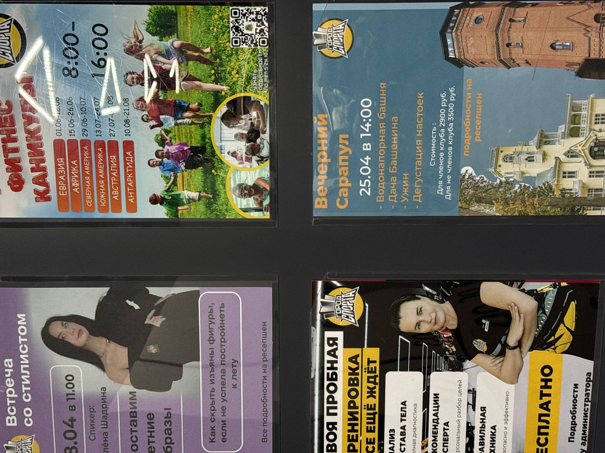 The image shows a bulletin board with four flyers advertising different events in the city of Votkinsk, Russia.
The upper-left flyer advertises "Fitness Vacations" to different locations such as Eurasia, Africa, North America, South America, Australia, and Antarctica, with scheduled dates and times from June to August. The upper-right flyer advertises an evening in Sarapul on April 25th at 2:00 PM, including a visit to the water tower and Bashenin's dacha with dinner and a tasting of infusions. Prices for club members and non-club members are listed. The lower-left flyer promotes a consultation with stylist Alyona Shadrina on April 3rd at 11:00 AM, with tips on how to hide flaws with clothes. The lower-right flyer advertises a free trial workout with body composition analysis, expert recommendations, and correct technique.