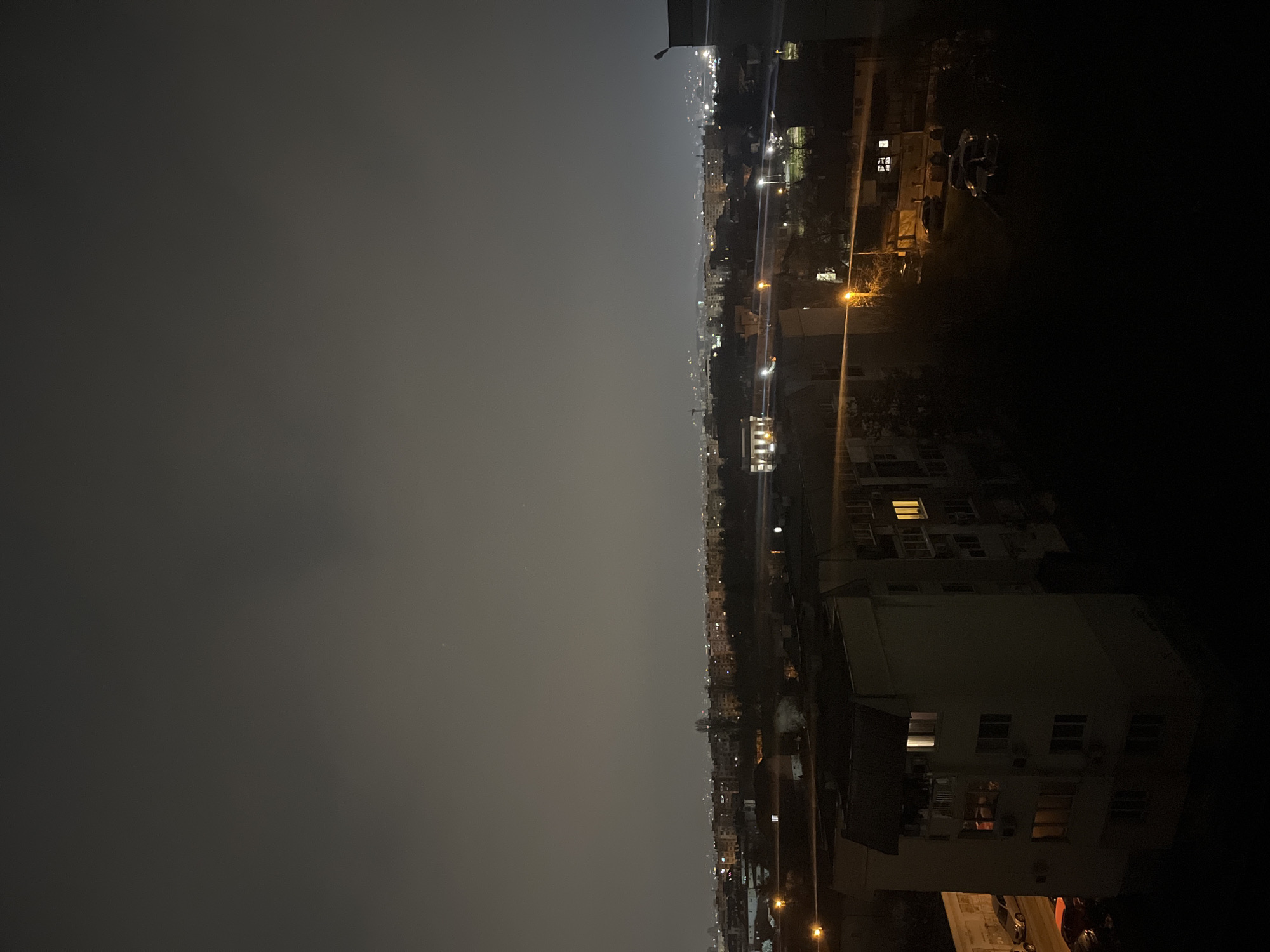The image depicts a nighttime view of a city skyline, specifically Sofia, Bulgaria. The right side of the image reveals residential buildings, illuminated by streetlights and lights from individual apartments. There are several cars parked near the building. The sky is overcast and dark.