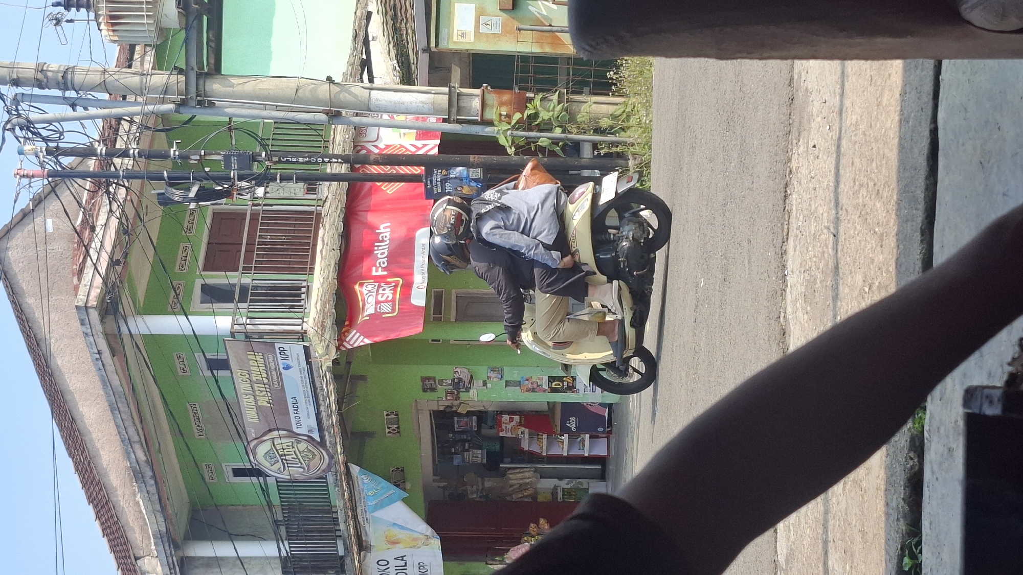 In the city of Cilaku, Indonesia, a beige scooter transports two individuals. The rider, a man wearing a black jacket and beige pants, is seated in front. Behind him, a woman with a gray shirt and headscarf is the passenger. Both are wearing helmets for safety. The scooter is in motion on an asphalt road, passing by a two-story building with a green facade. A banner above the ground floor of the building displays "KUALITAS TINGGI PASTI JUARA!" and "TOKO FADILA". The scene is illuminated by daylight under a clear blue sky. Electrical poles with numerous wires are visible near the building. The foreground shows a blurred view of a person's arm and a table.