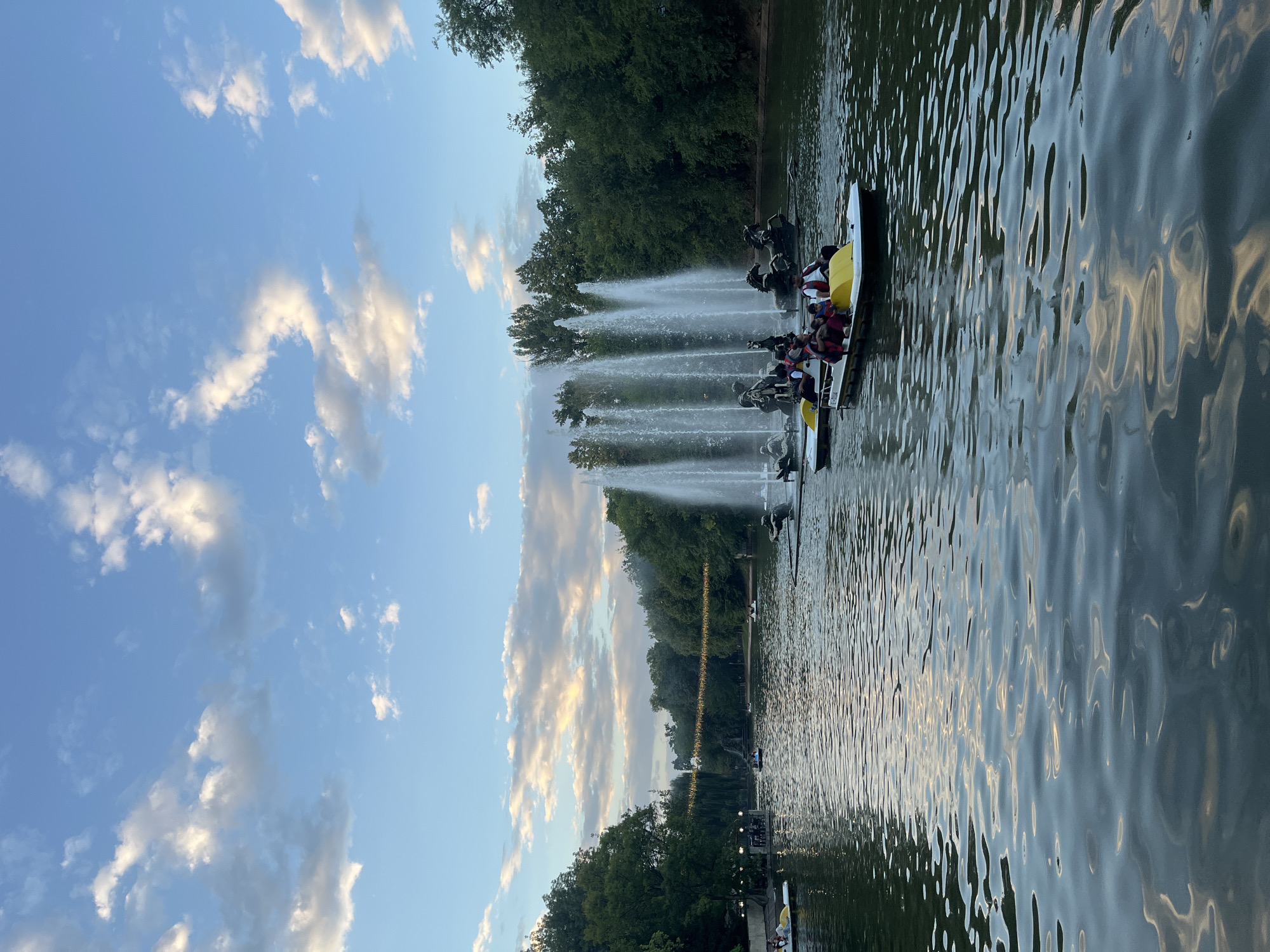 The image depicts a scenic view of a park with a large body of water, likely a lake or pond, in Almaty, Kazakhstan. The sky is a clear blue with scattered white clouds, suggesting it is daytime, possibly late afternoon or early evening given the lighting. Dominating the center of the water is a large fountain with multiple jets of water shooting upwards. In front of the fountain, there are several pedal boats on the water. Some of these boats are occupied by people enjoying a leisurely ride. The boats are white with yellow accents. Surrounding the water is lush green foliage and trees, indicating a park setting. In the distance, there are more trees and what appears to be some structures and possibly decorative lights strung between trees. The water surface has gentle ripples and reflects the sky and surrounding environment. The overall atmosphere is peaceful and recreational.