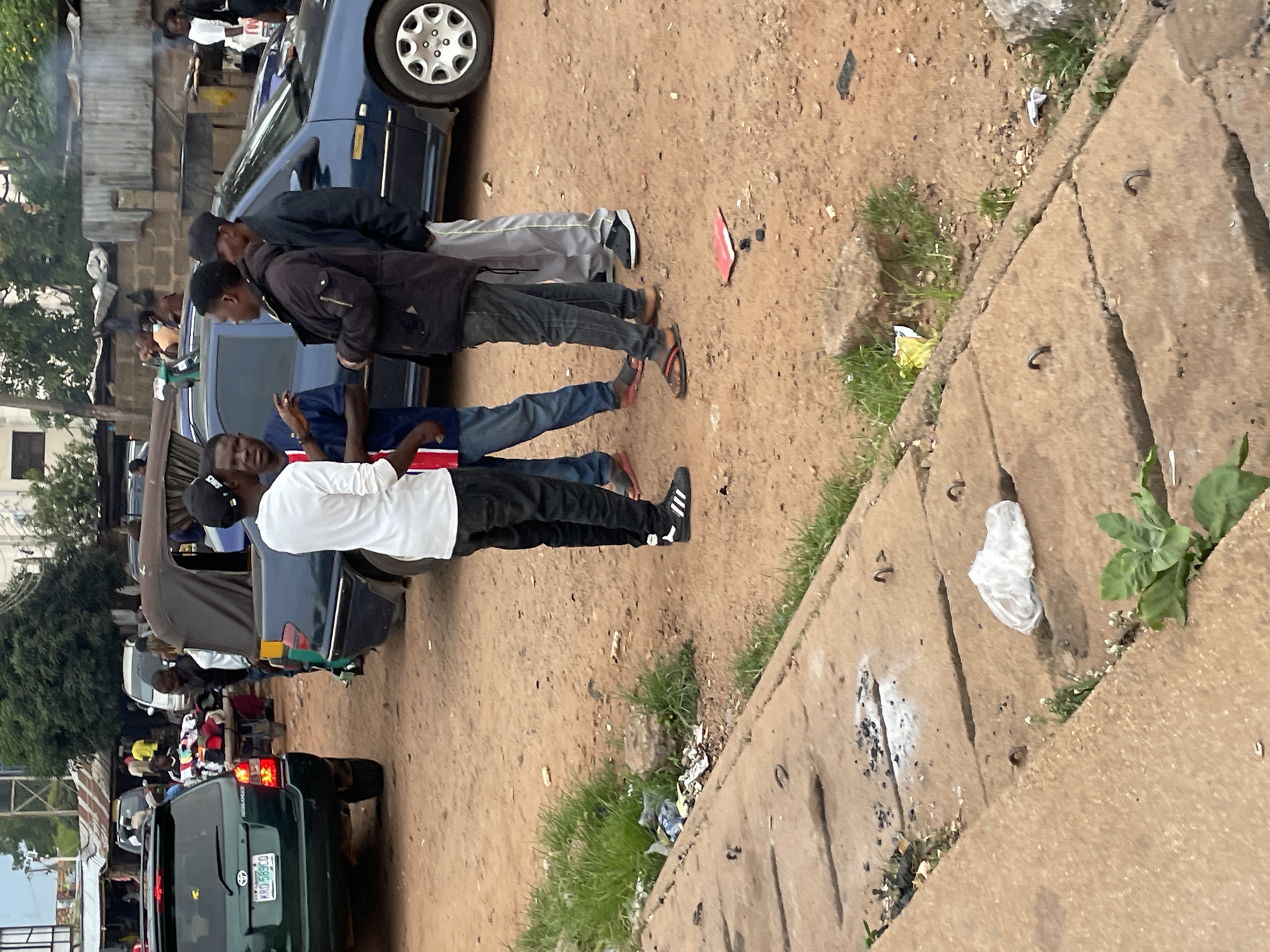 In Jos, Nigeria, a group of four young men are gathered on a dusty, uneven ground, possibly near a roadside market or bus station. The man on the left, wearing a white long-sleeved shirt and black pants, appears to be speaking with the man next to him, who is wearing a blue t-shirt with a red and white stripe. The third man, in a black jacket and dark jeans, stands facing away from the viewer, while the fourth man, in a black cap and gray pants, stands with his back to the camera.
The environment suggests a bustling urban setting with a mix of vehicles and structures. A dark blue sedan is parked behind the group, its wheel visible. To the left, the rear of a green SUV with a visible license plate (KRD 589CO) is present. In the background, a dark-colored open-top vehicle, possibly a commercial bus or a modified van, is partially visible. The ground is a mixture of red earth, with patches of dry grass and scattered debris, including a red object that might be a piece of paper or plastic, and a white plastic bag. Concrete slabs with metal hooks are visible in the foreground, suggesting a drainage or sidewalk area. In the distance, corrugated metal roofing and brick walls indicate buildings, and smoke or steam is rising from behind them, hinting at cooking or other activities. The overall scene depicts a moment of informal gathering or interaction in a vibrant, somewhat unkempt urban environment.