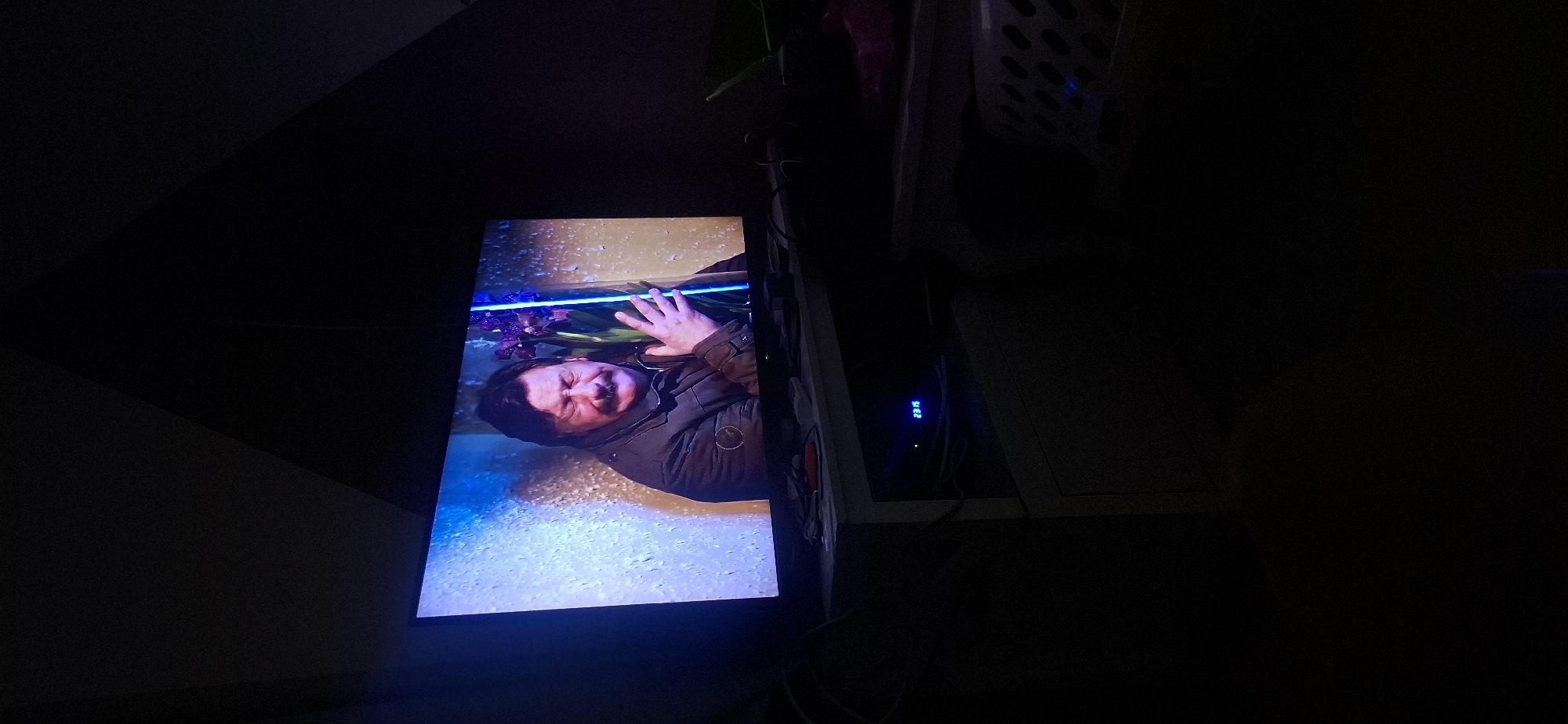The image shows a television screen displaying a man with his eyes closed, hugging flowers. The scene appears to be indoors, with the room dimly lit. The television is placed on a shelf, and some electronic devices are visible below it. A digital clock on one of the devices shows the time as 23:15. The background of the image is dark, suggesting it is nighttime. The provided context states the image is from Yerocham, Israel, but there are no visual cues in the image to confirm this location.