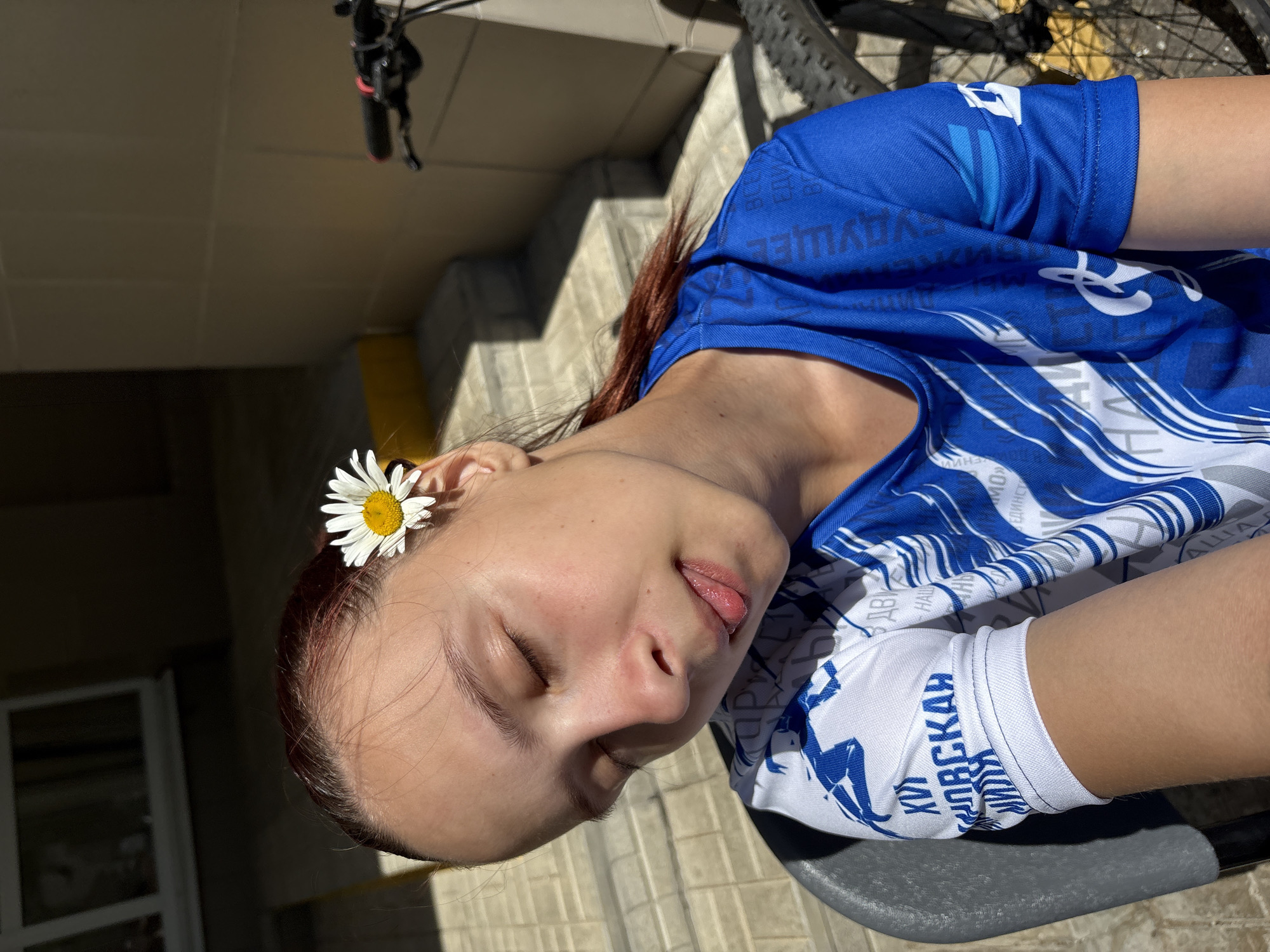 A young woman with her eyes closed, a daisy tucked behind her right ear, and her tongue slightly protruding, is shown in a close-up shot. She is wearing a blue and white patterned t-shirt. The setting appears to be outdoors, with a tiled pathway and steps visible behind her. The lighting suggests it is daytime, with sunlight casting shadows. A bicycle is partially visible to the right of the frame. This scene is located in the city of Chaikovsky, Russia.