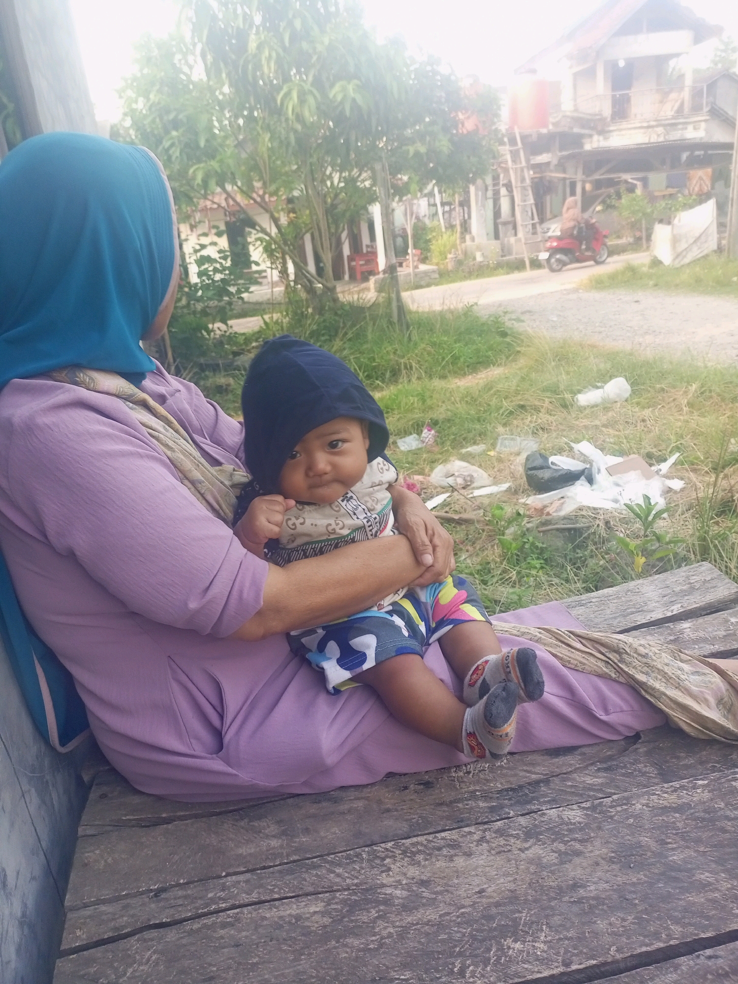 In the city of Jawilan, Indonesia, a woman wearing a turquoise hijab and a lavender dress is seated on a wooden plank bench. She is holding a baby wearing a dark blue hood and a patterned onesie with colorful shorts. The baby is facing the camera, and their feet are visible, clad in grey socks with orange and black accents. The background shows a rural scene with a modest house, a ladder leaning against a water tank, a red scooter, and overgrown grass with scattered debris. The lighting suggests it is daytime, possibly late afternoon due to the soft, diffused light. The overall setting appears to be outdoors, in a residential area.
