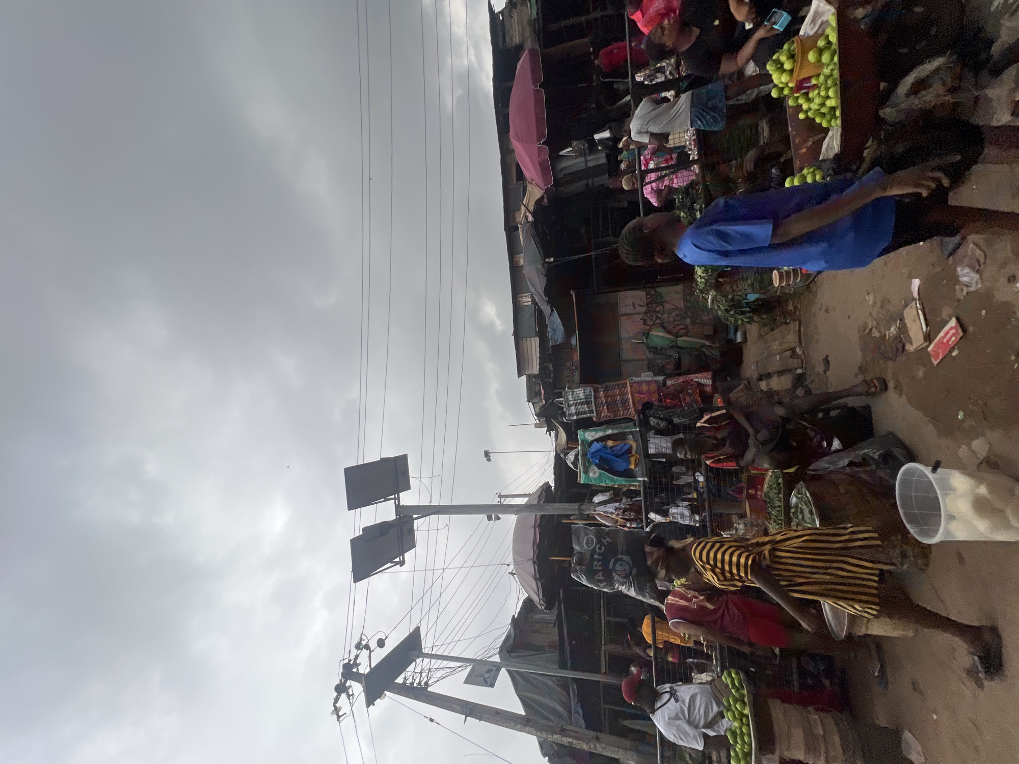 The image depicts a busy outdoor market in Nkpor, Nigeria, under an overcast sky. Several stalls with corrugated metal roofs are visible, displaying various goods including what appear to be textiles and produce. In the foreground, people are engaged in market activities. A person in a yellow and black striped dress walks past a stall piled high with green fruits. Another person, wearing a white shirt and a red cap, stands near a display of green fruits. The market appears to be crowded with people, some of whom are vendors and others are shoppers. Power lines and utility poles are prominent in the background. The ground is dirt and appears somewhat wet, suggesting recent rain or general market conditions.