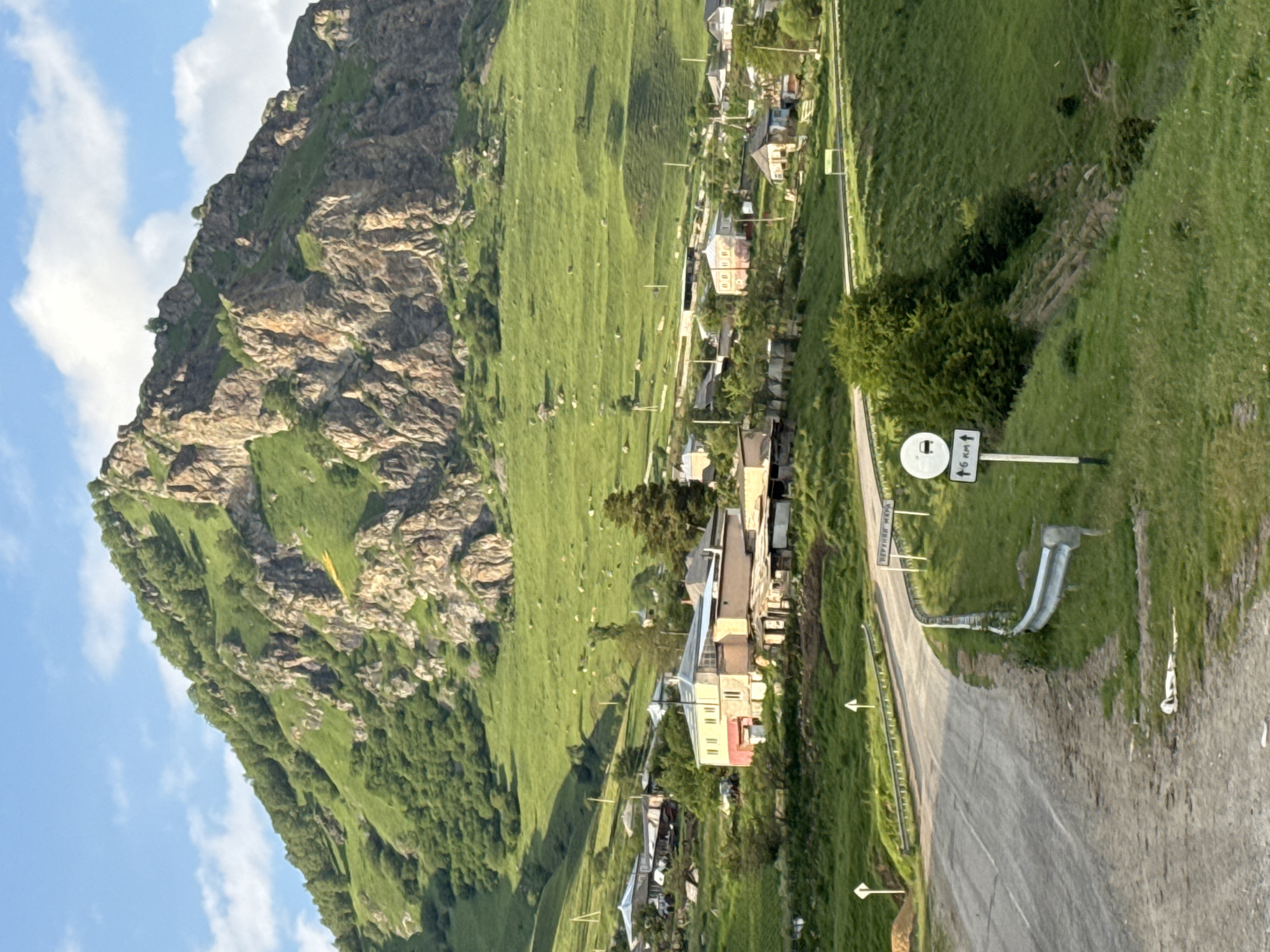 A paraglider with a yellow wing is visible against the backdrop of a steep, rocky mountainside covered in lush green grass. The mountain slopes gently down to a village with several houses nestled amongst the greenery. A paved road winds through the village, with some houses situated close to the road. Several white directional signs are posted along the road. One sign features a circle with an arrow pointing right and the text "6 км" which translates to "6 km". Another sign displays Russian text, which appears to be "НИЖНЯЯ МАРА" (Nizhnyaya Mara), indicating the village's name. The sky is blue with scattered white clouds. The overall scene suggests a bright, sunny day in a mountainous region.