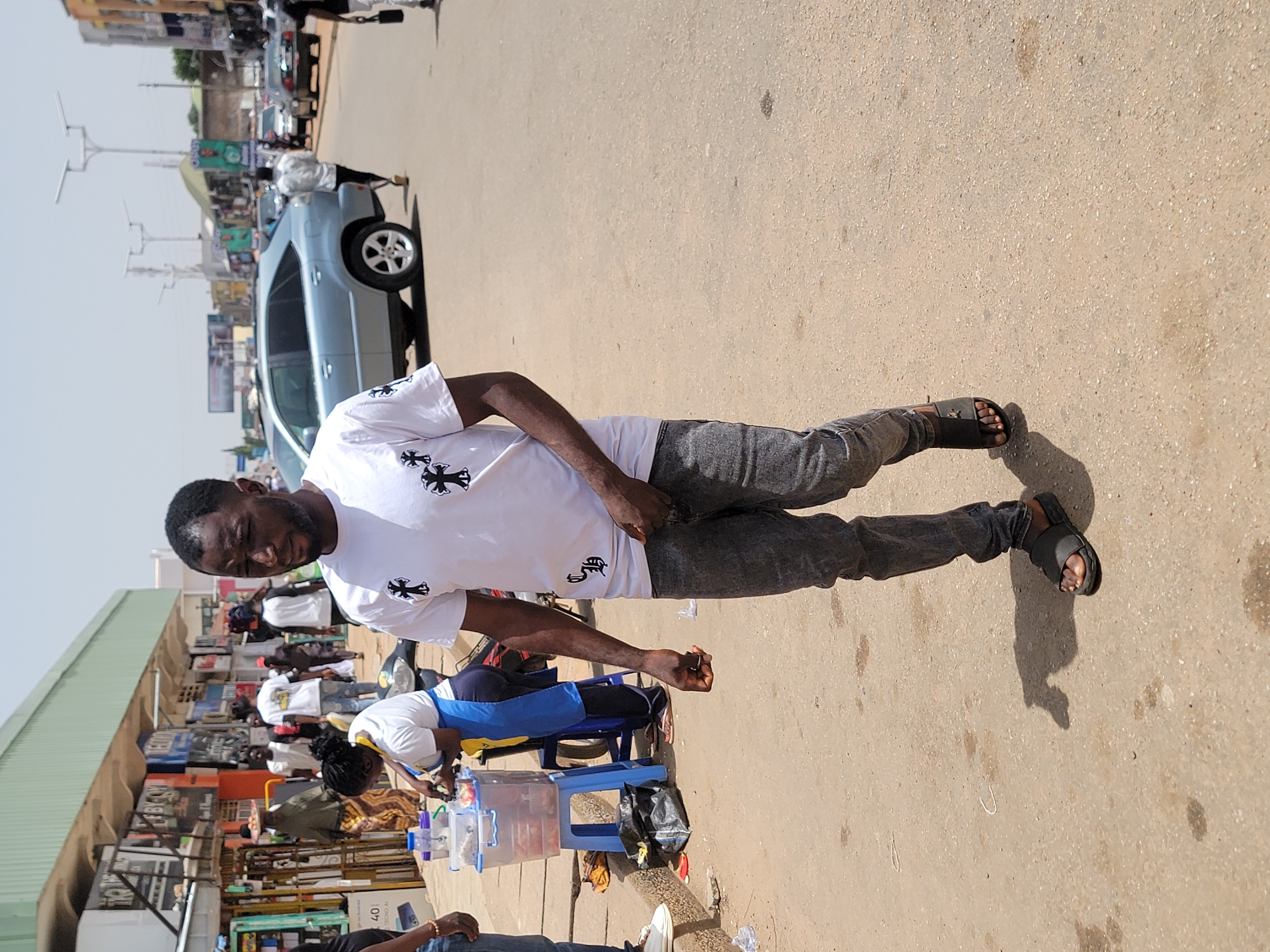 A man is standing on a street in Jos, Nigeria. He is wearing a white t-shirt with a black cross pattern, gray jeans, and black sandals. The street appears to be in a commercial area with shops on one side. There is a car parked nearby. There are other people on the street, including one seated behind a table with a water dispenser and cups. The overall scene suggests a bustling outdoor market or commercial district in Jos.