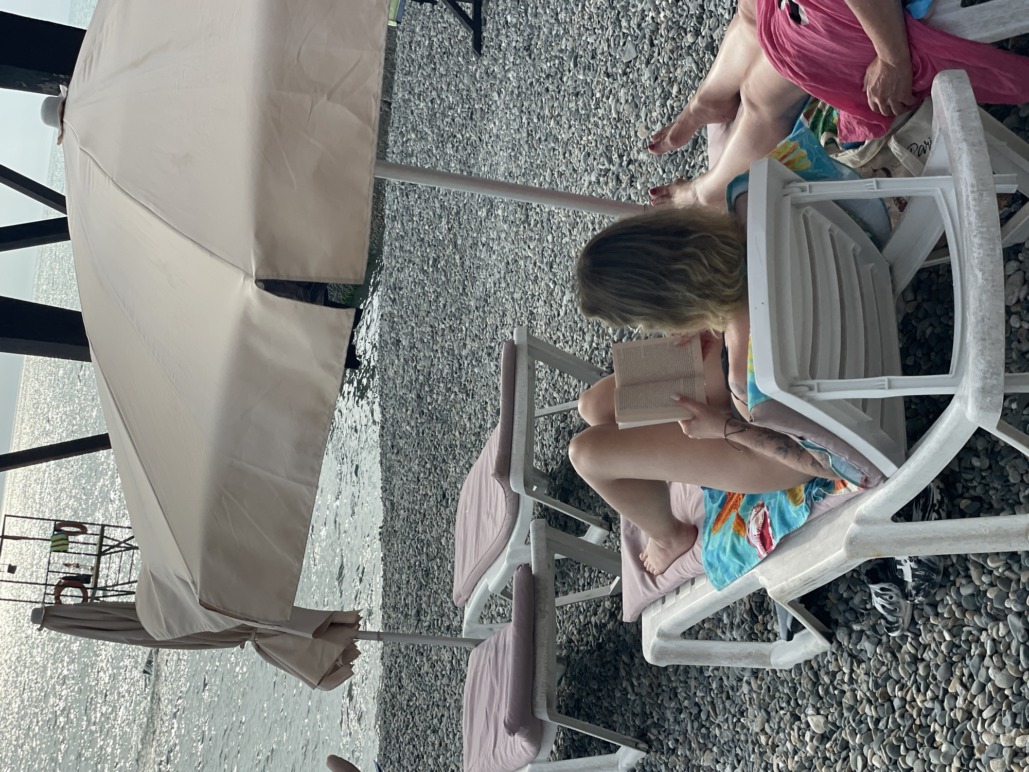 Two people are relaxing on lounge chairs on a pebble beach next to the sea. One person, with blonde hair, is reading a book while sitting in a white plastic lounge chair. They are wearing a colorful swimsuit. Another person, wearing a bright pink sarong, is sitting in a lounge chair next to them, with their legs visible. The sea is calm and reflects the light. A large beach umbrella provides shade. In the background, a lifeguard tower with lifebuoys is visible. The scene suggests a leisurely day at the beach, likely during daylight hours. The overall mood is relaxed and peaceful. The location is identified as Sochi, Russia.