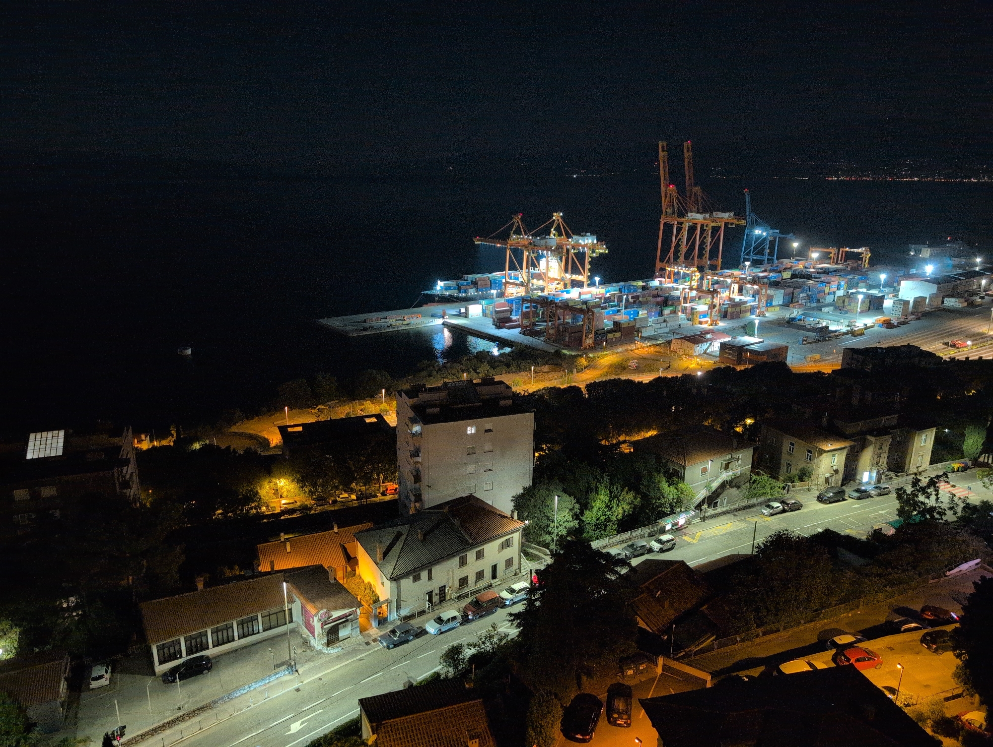 This aerial night photograph captures a section of the city of Grad Rijeka, Croatia, with a prominent focus on a port area and surrounding urban landscape. The scene is illuminated by artificial lighting from the port facilities, streetlights, and building interiors.
In the upper portion of the image, a large, active port is visible, characterized by numerous shipping containers stacked high and a series of towering cranes, several of which are illuminated. The dark expanse of the sea stretches beyond the port. The distant shoreline is dotted with lights, suggesting other urban areas or settlements.
Closer to the foreground and extending towards the bottom of the frame, the residential and commercial areas of the city are depicted. These areas feature multi-story buildings, smaller houses with red-tiled roofs, and roadways. Several vehicles are visible on the streets, including cars parked along the roadside and in driveways. The trees and vegetation are silhouetted against the artificial light. The overall impression is of a bustling port city at night, with the industrial activity of the harbor juxtaposed against the quieter residential zones.
