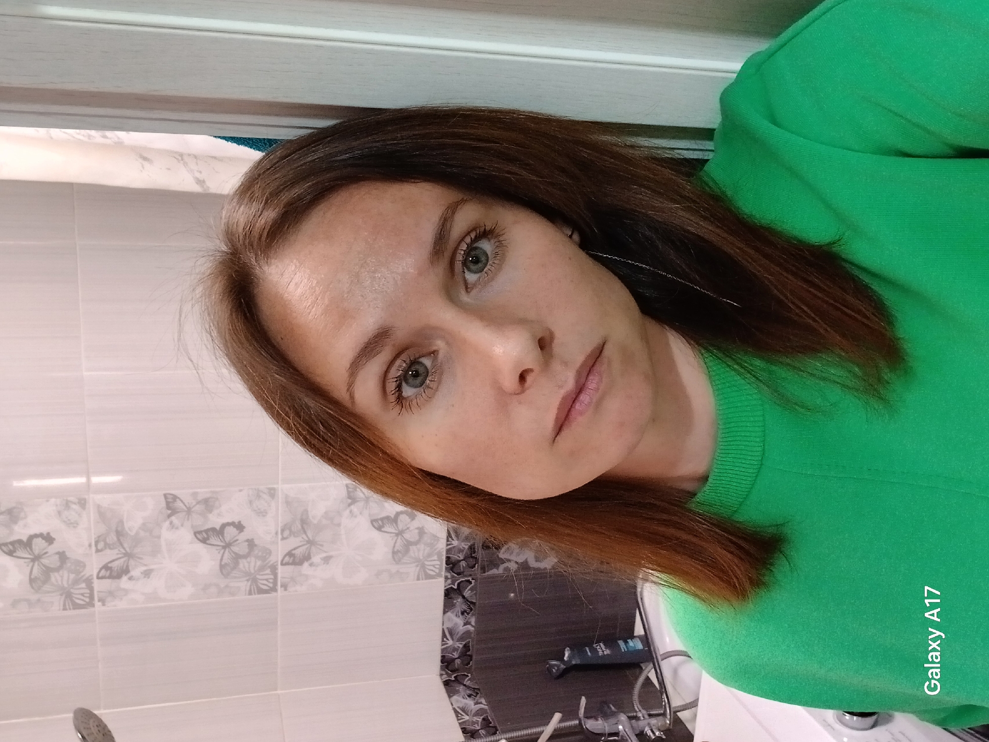 A woman with light skin, green eyes, and auburn-brown hair is facing the camera. She is wearing a green sweater. The background consists of a tiled wall with a butterfly pattern, a sink, and bathroom fixtures. The image appears to have been taken in a bathroom, but there is no information available to confirm that the location is in Yekaterinburg, Russia.