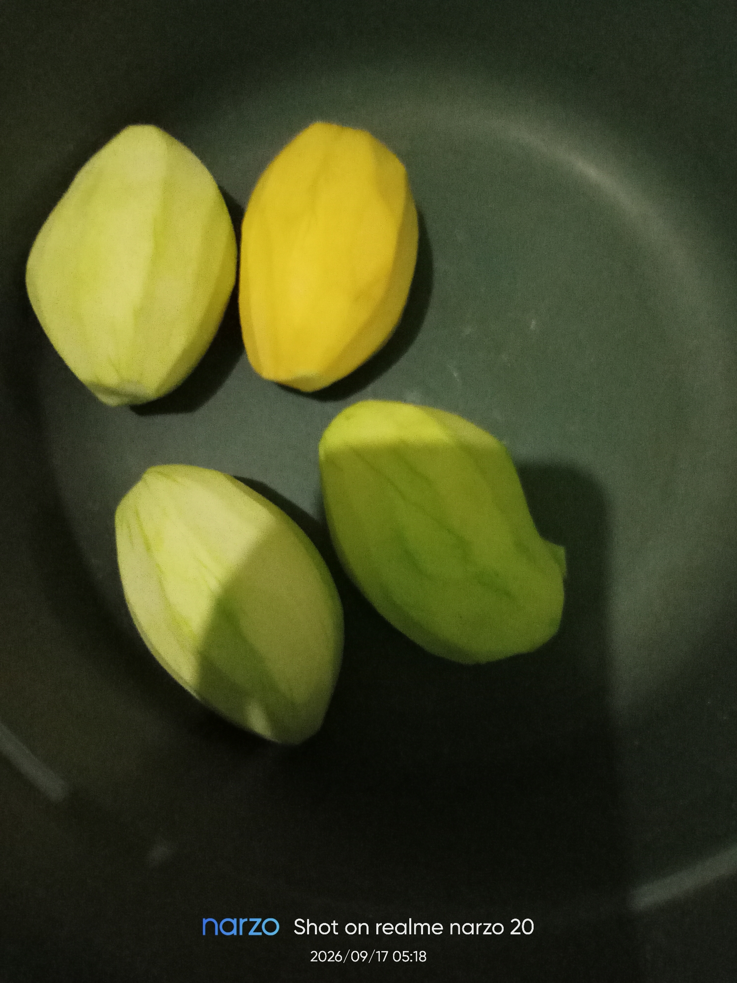 The image shows four peeled mangoes arranged in a circular pattern on a dark green, textured surface, possibly a pan or bowl. Three of the mangoes are light green, while one is bright yellow. Shadows are cast by the mangoes, suggesting directional lighting from above and to the left. The background is dark and out of focus. At the bottom right corner of the image, a watermark reads "narzo Shot on realme narzo 20" and "2026/09/17 05:18". The image does not contain any information about the city of Karawang, Indonesia.