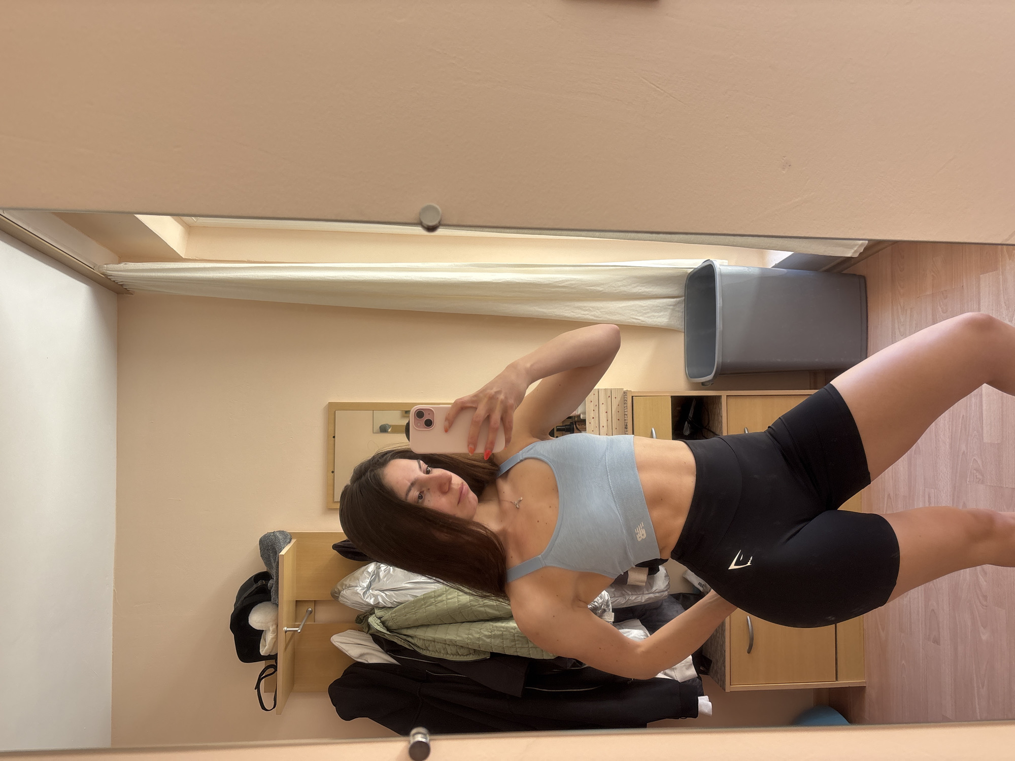 A woman takes a selfie in front of a mirror. The reflection shows her wearing a blue sports bra with a white New Balance logo and black workout shorts with a white Gymshark logo. She is holding a pink iPhone. The background includes a clothes rack with coats, a dresser, a gray trash bin, and a window with a beige curtain. The setting appears to be a room in a residential building with beige walls. There is no definitive evidence indicating the location is Varna, Bulgaria.