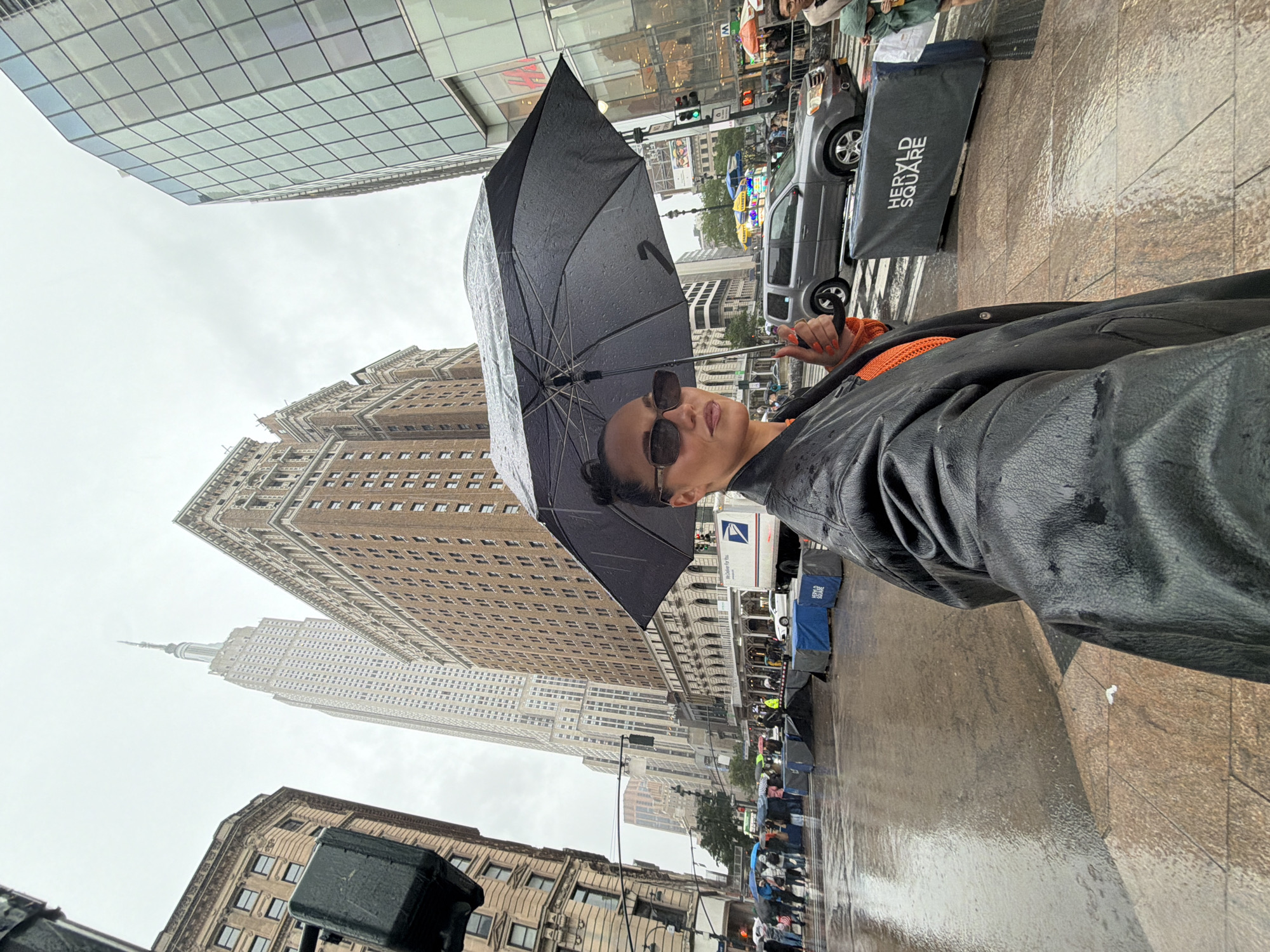 The image is a low-angle shot of a person holding a black umbrella in the rain in New York City. The Empire State Building is visible in the background. Other surrounding buildings are visible on the left of the image. There is a street scene on the right of the image showing pedestrian traffic, cars, traffic lights and a tent, with the words 'Herald Square' on its side. The ground is wet. The person is wearing sunglasses and a black leather jacket, with an orange top underneath.